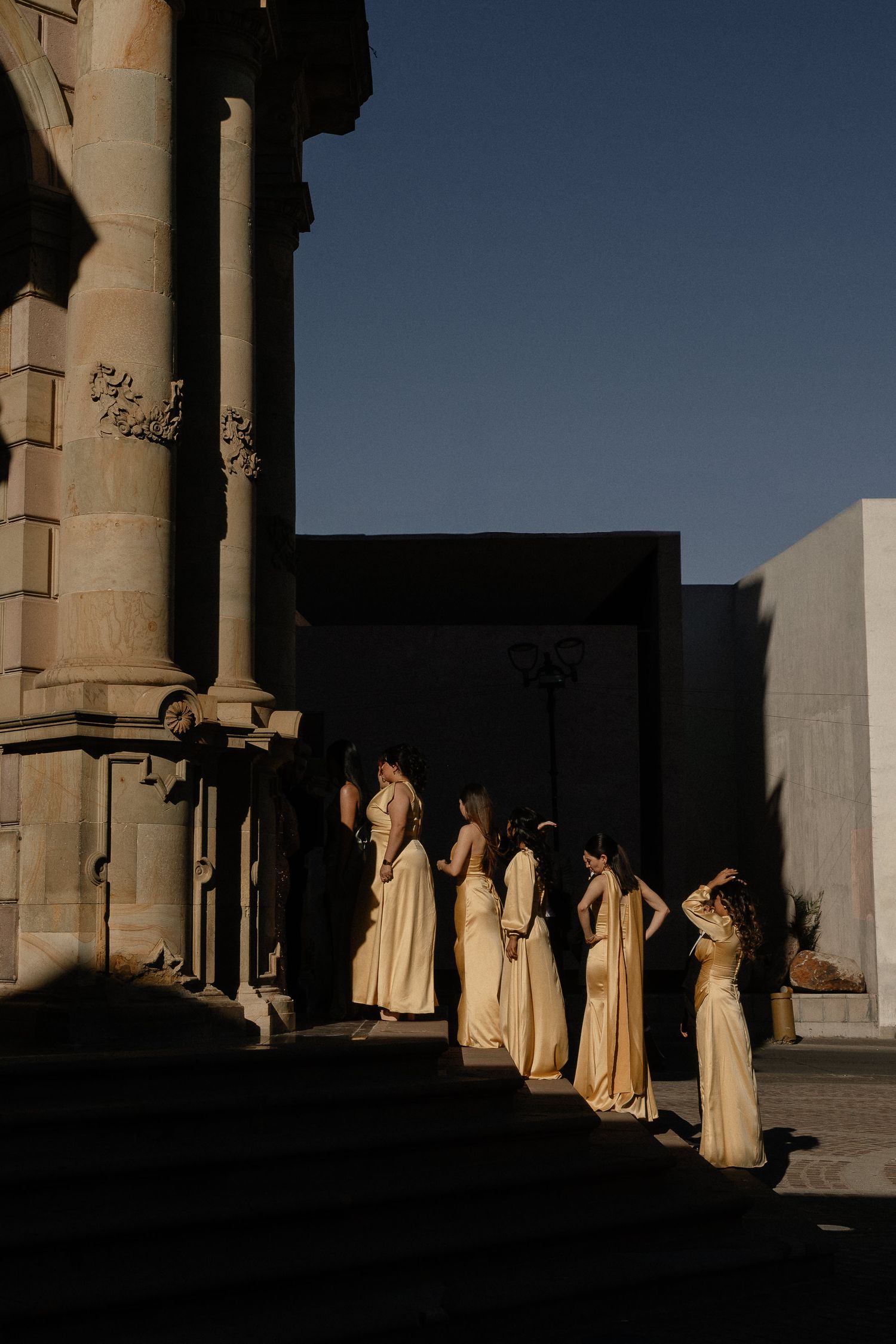 Figures in cream-colored robes stand between stone columns casting dramatic shadows in evening light.