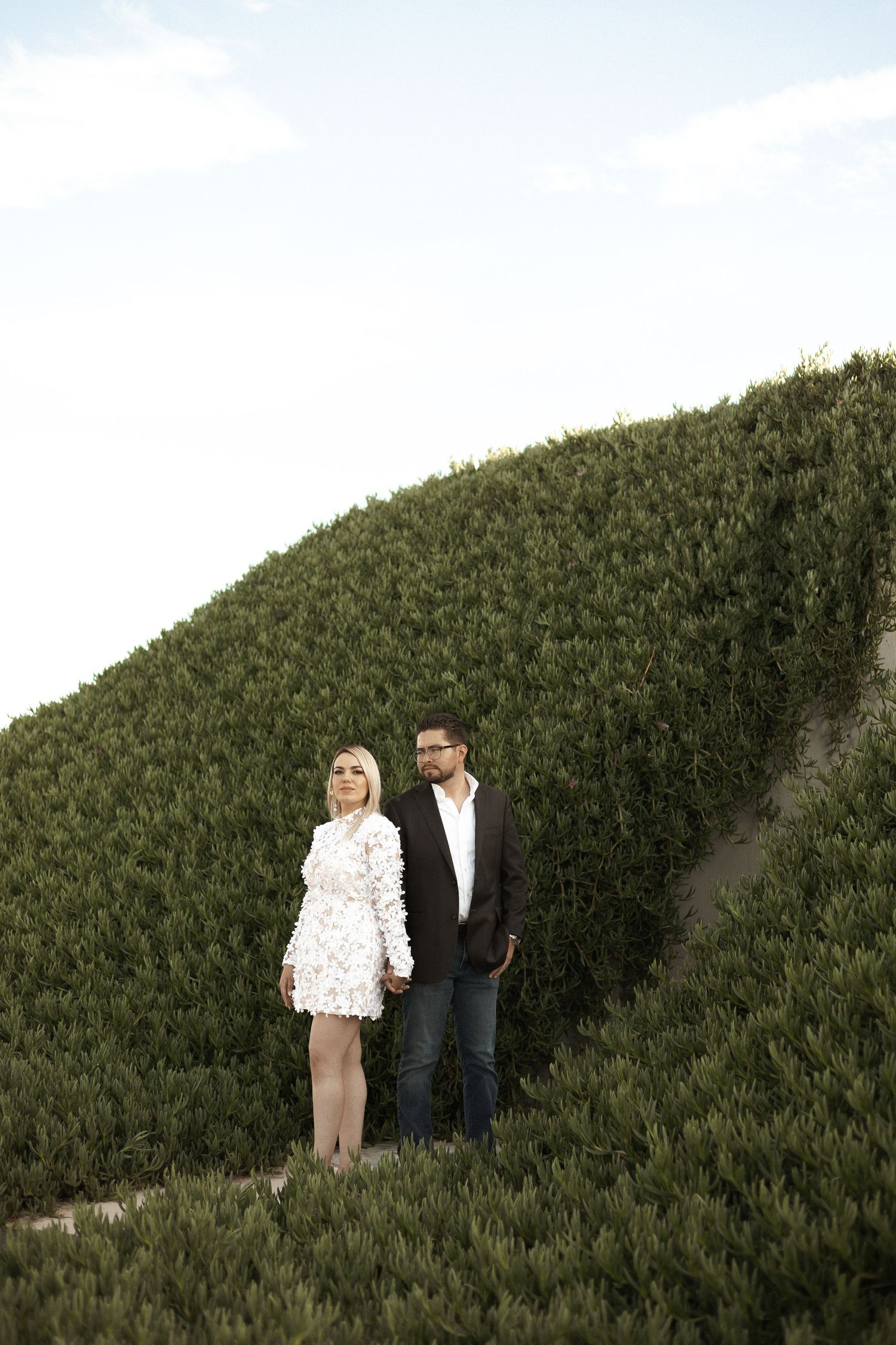 Two people in formal attire stand together against a tall green hedge backdrop in an outdoor setting.