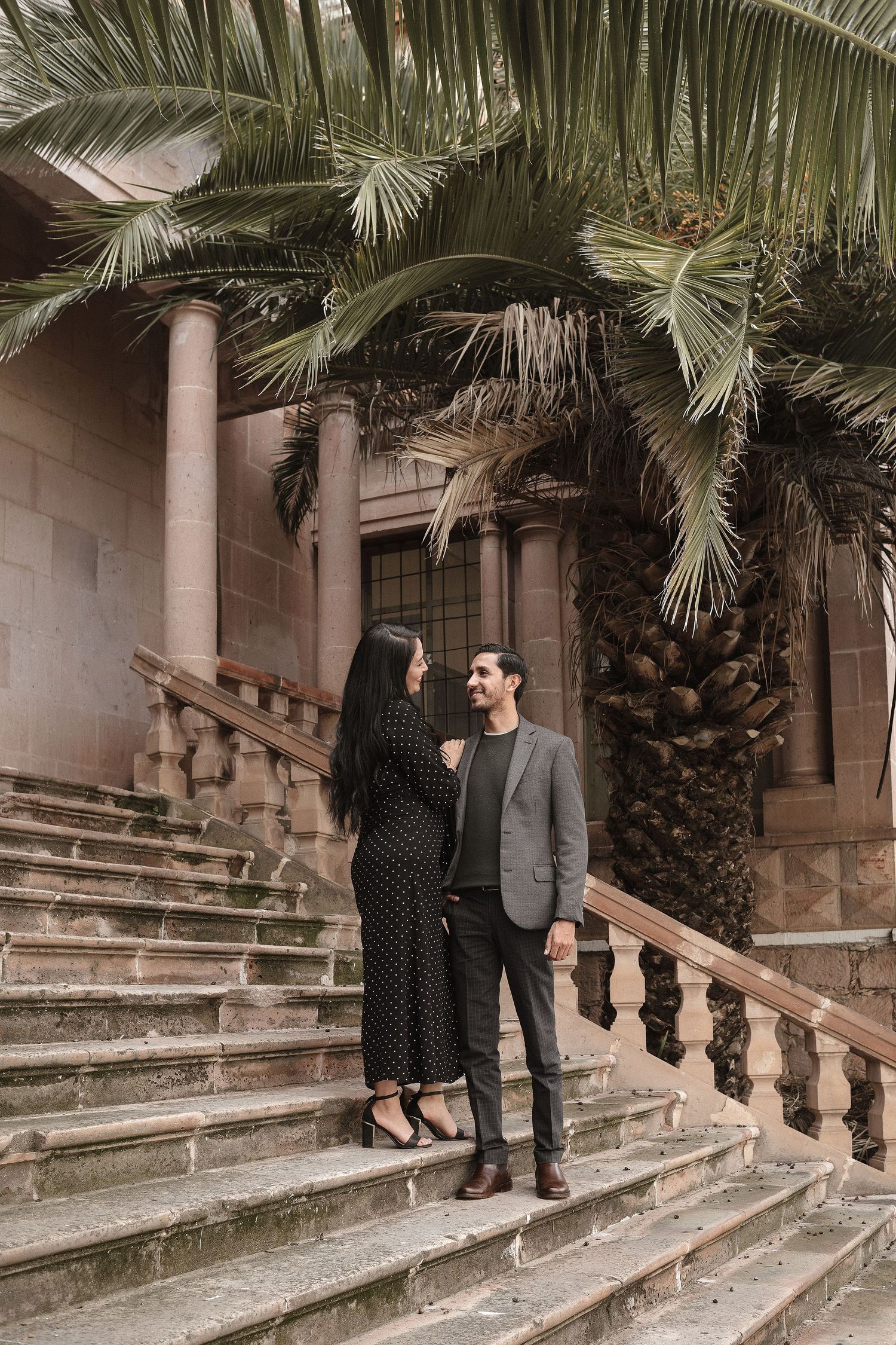 A romantic couple poses on stone steps surrounded by palm trees in an architectural setting.
