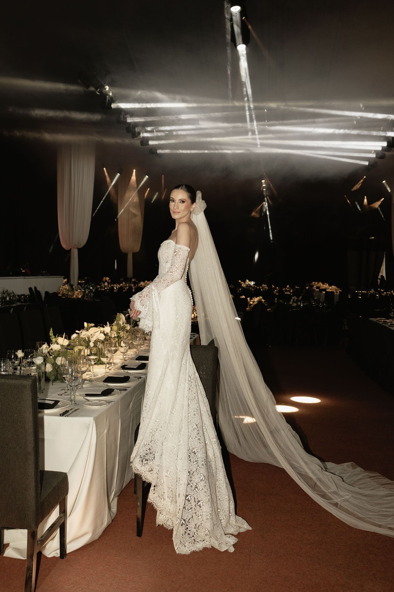 A bride in a fitted lace wedding gown with dramatic cathedral veil stands beside an elegant reception table setup.