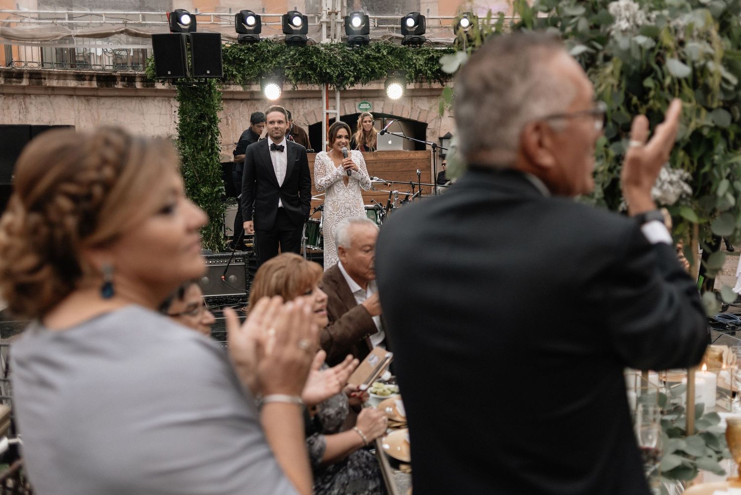 Wedding guests applaud during an outdoor reception celebration with string lights and greenery decorations.