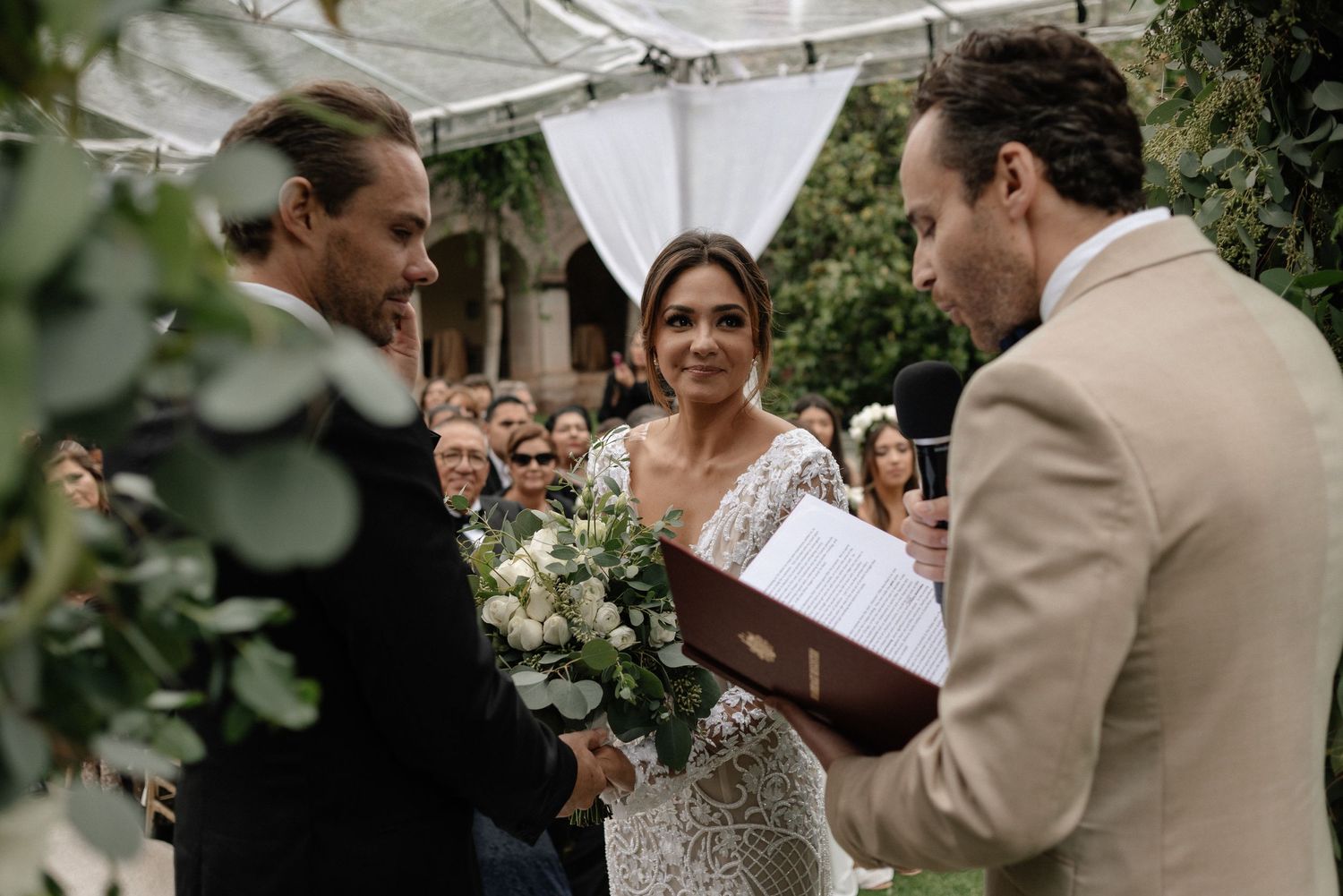 An intimate outdoor wedding ceremony takes place under a white canopy with lush greenery and white floral bouquets.