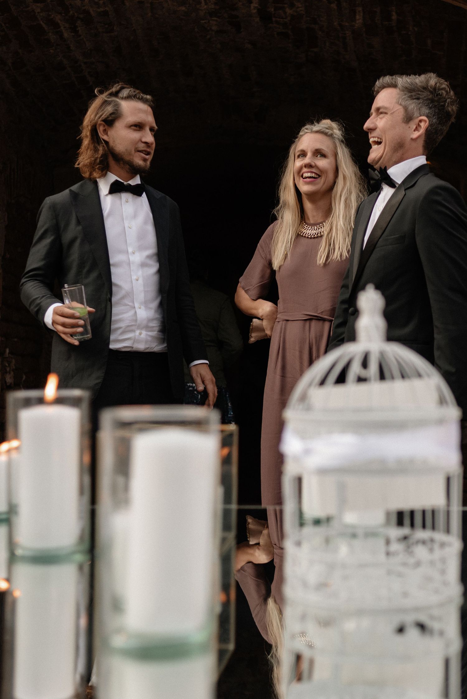 Wedding guests in formal attire laugh and socialize near decorative white candles and a birdcage at an evening celebration.