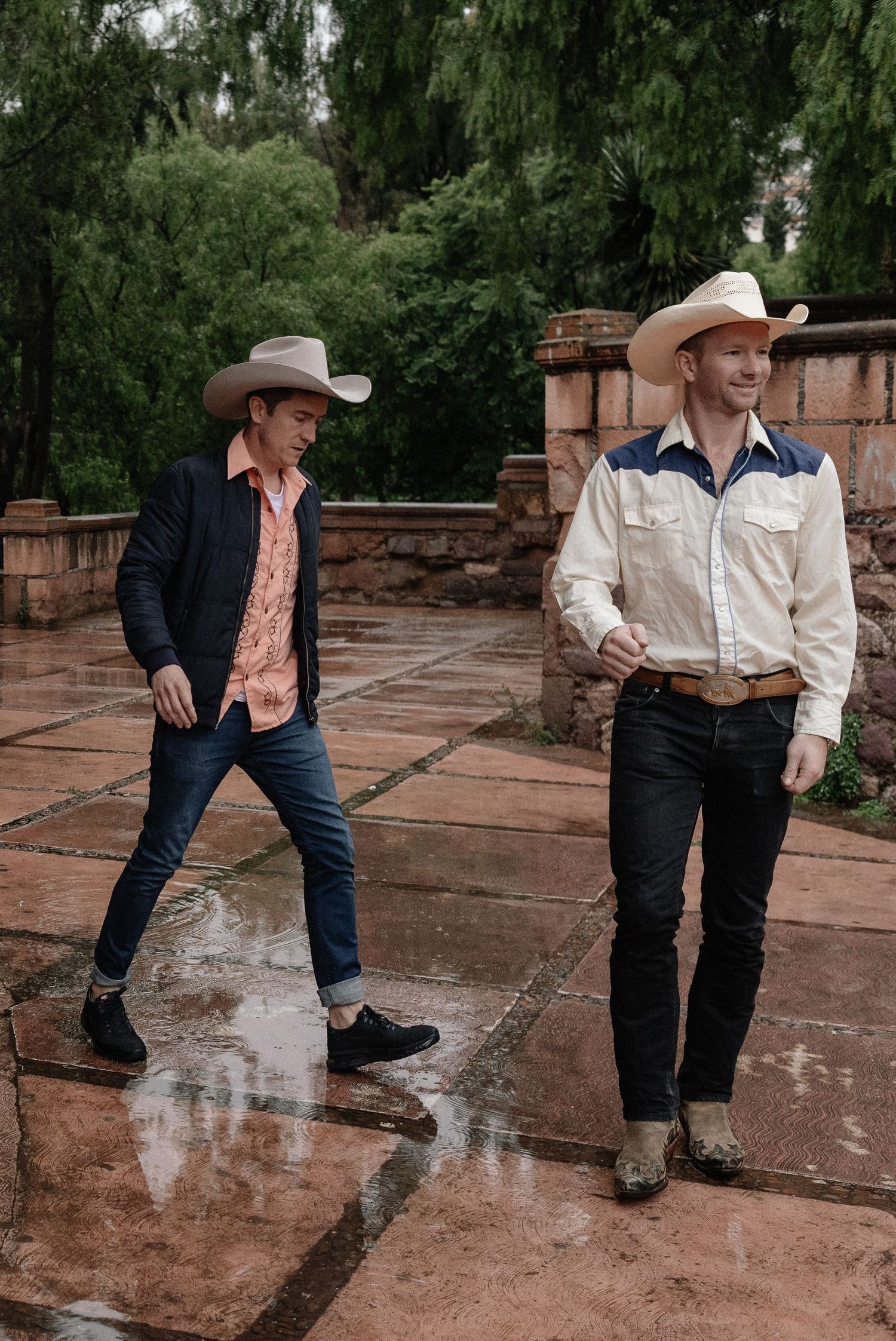 Two cowboys in denim and cowboy hats walk on a wet stone patio surrounded by green trees on a rainy day.