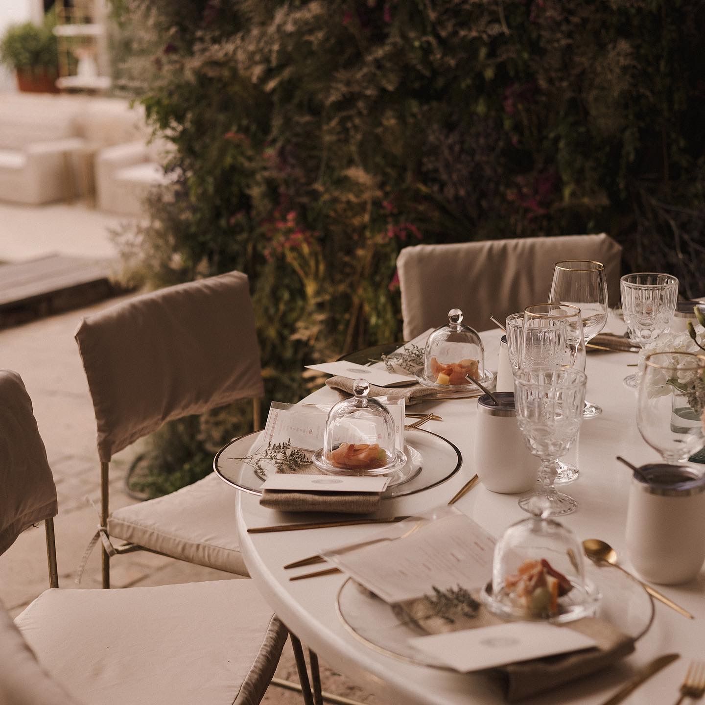 Elegant outdoor table setting with crystal glassware and covered plates in a romantic garden atmosphere.
