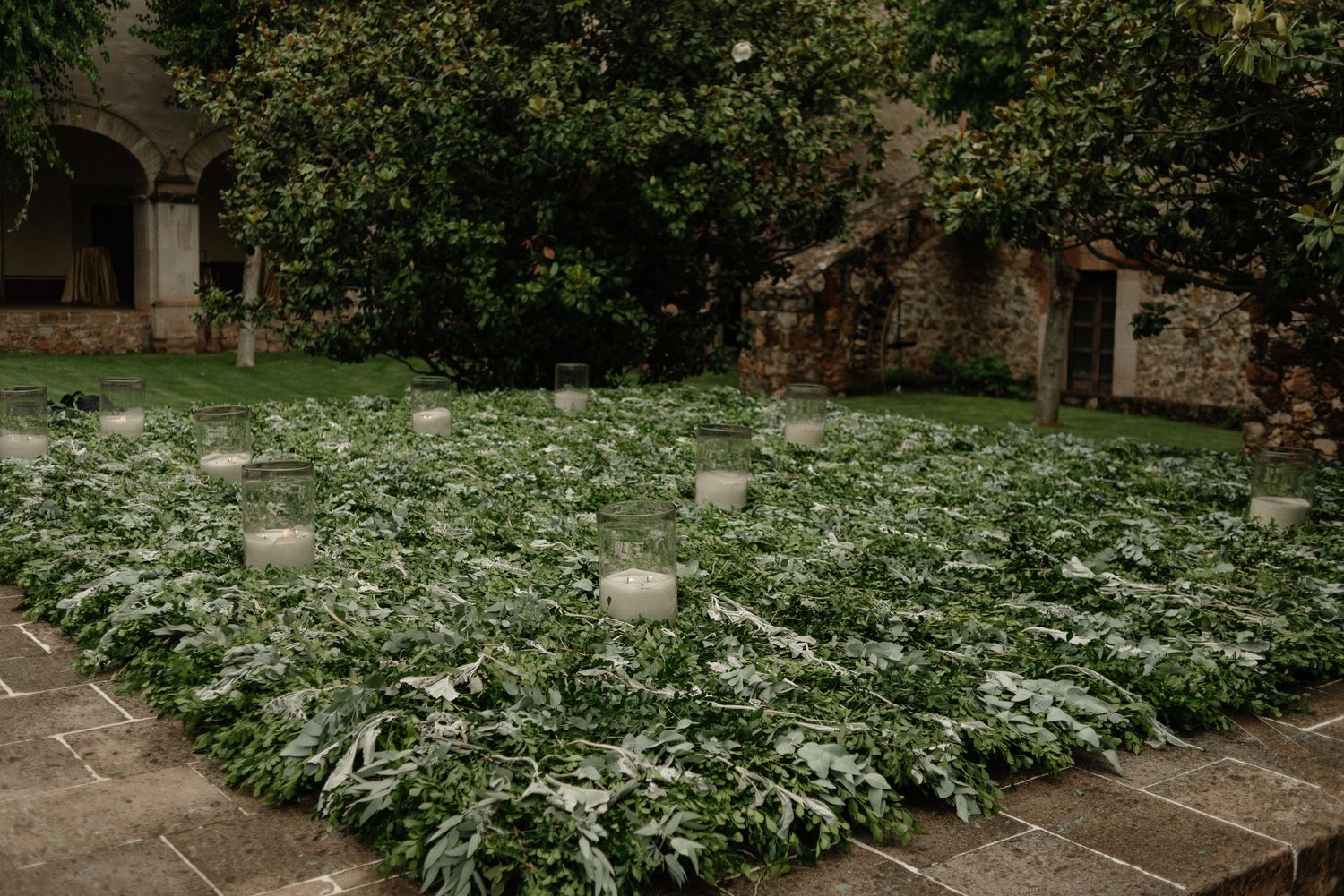 Lush green plants grow in a stone-walled garden courtyard with small markers scattered throughout the plot.