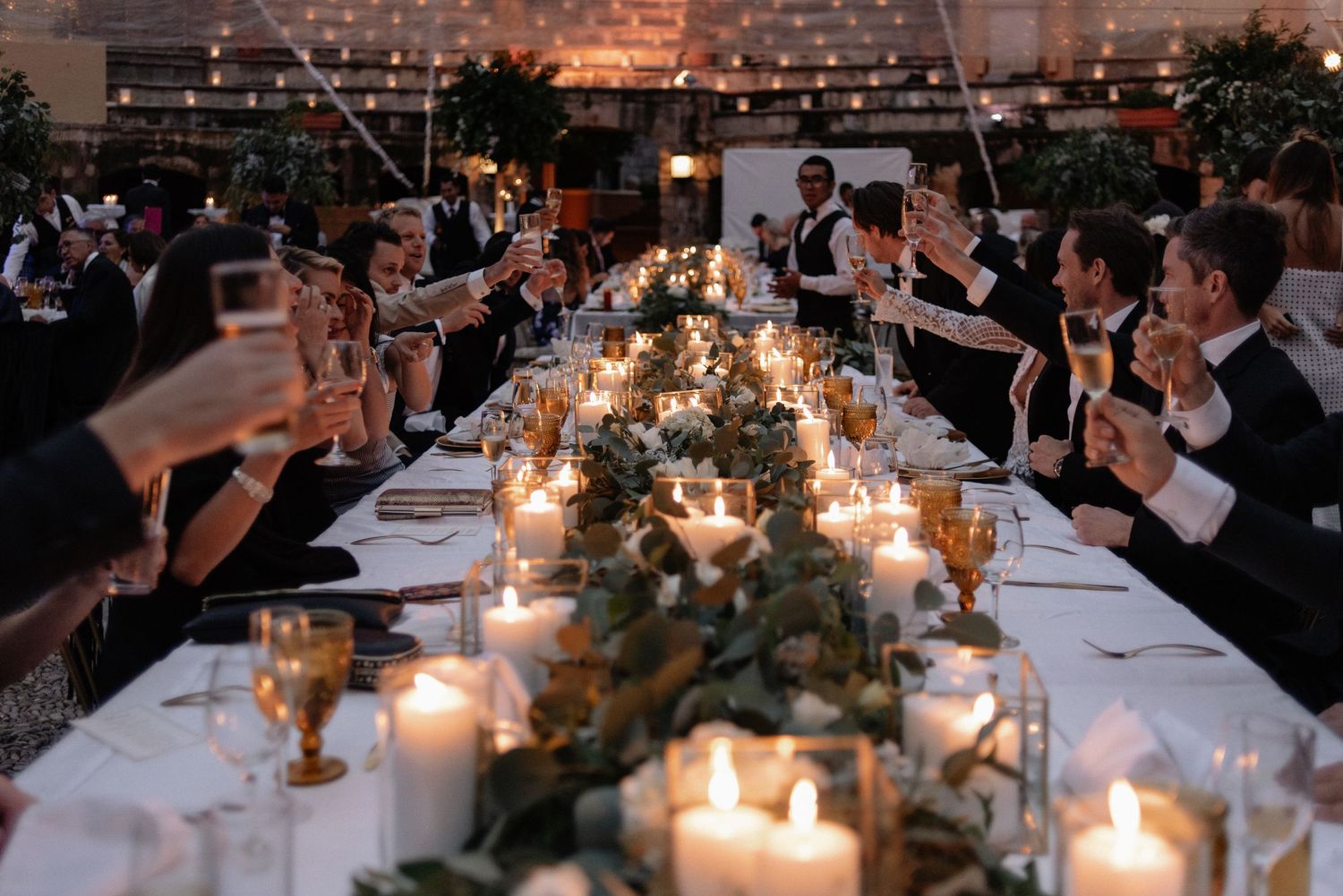 A candlelit wedding reception dinner table set with glowing candles and floral centerpieces as guests toast at twilight.