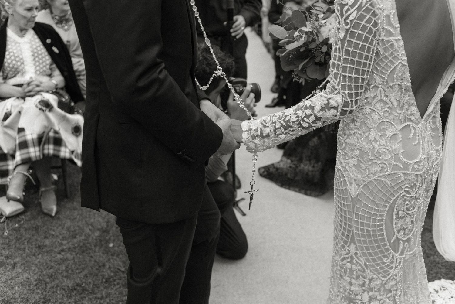 A close-up black and white photograph of newlyweds holding hands during an outdoor wedding ceremony.
