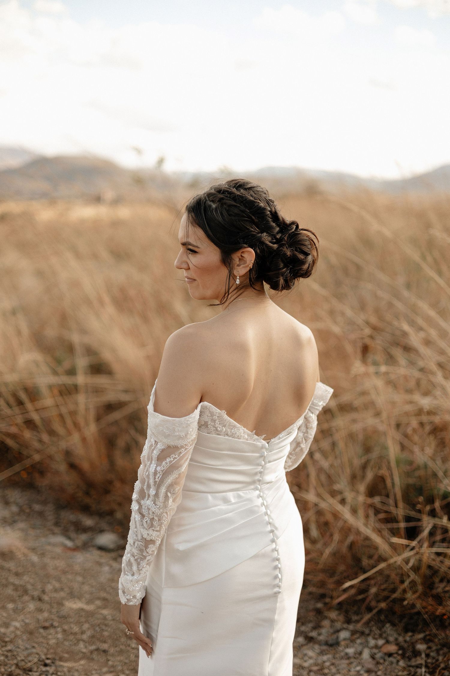A bride in a white lace wedding dress stands in a field of golden wheat against a mountainous backdrop at sunset.