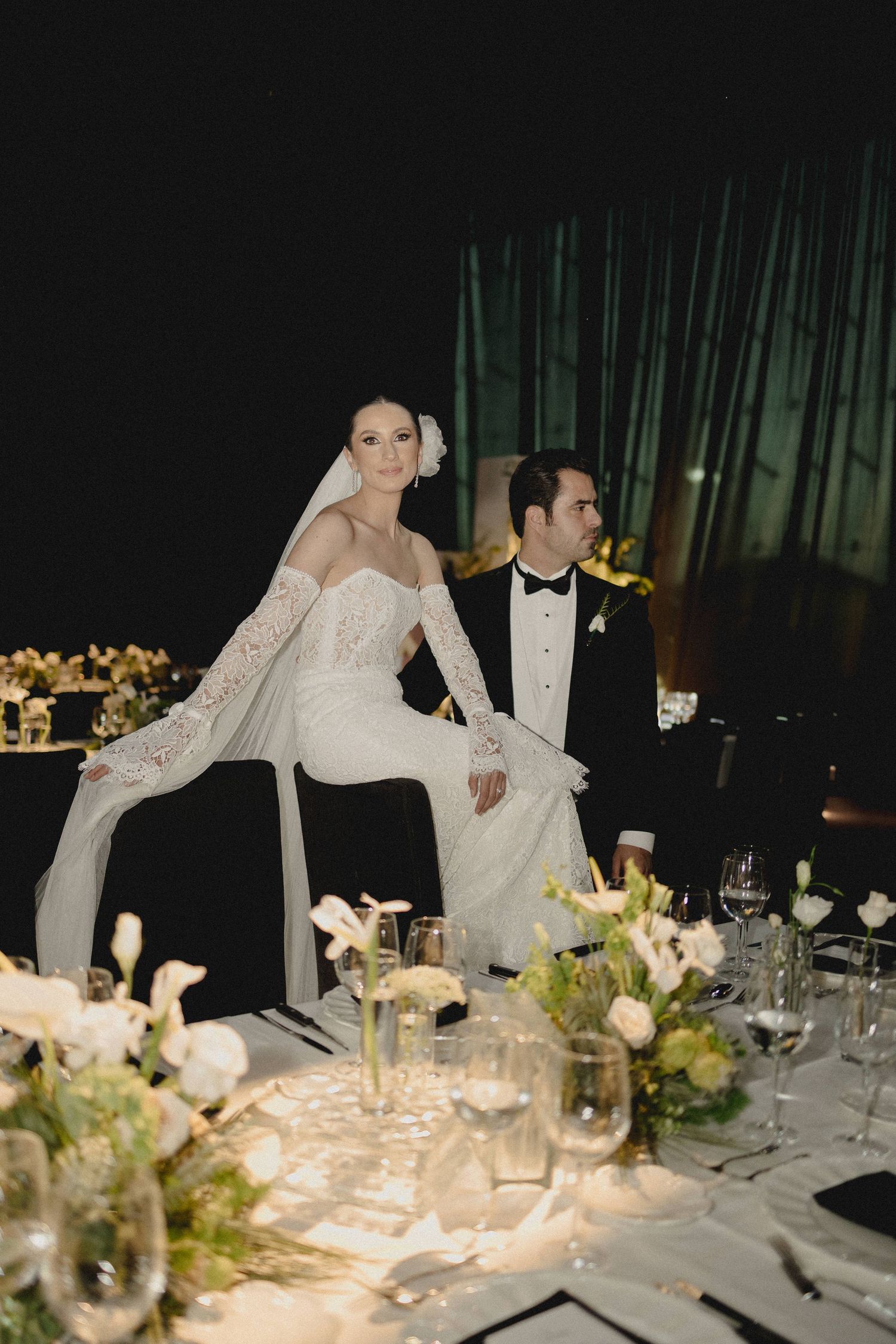 Elegant wedding reception table settings with white floral centerpieces and glowing candlelight in a dark venue.
