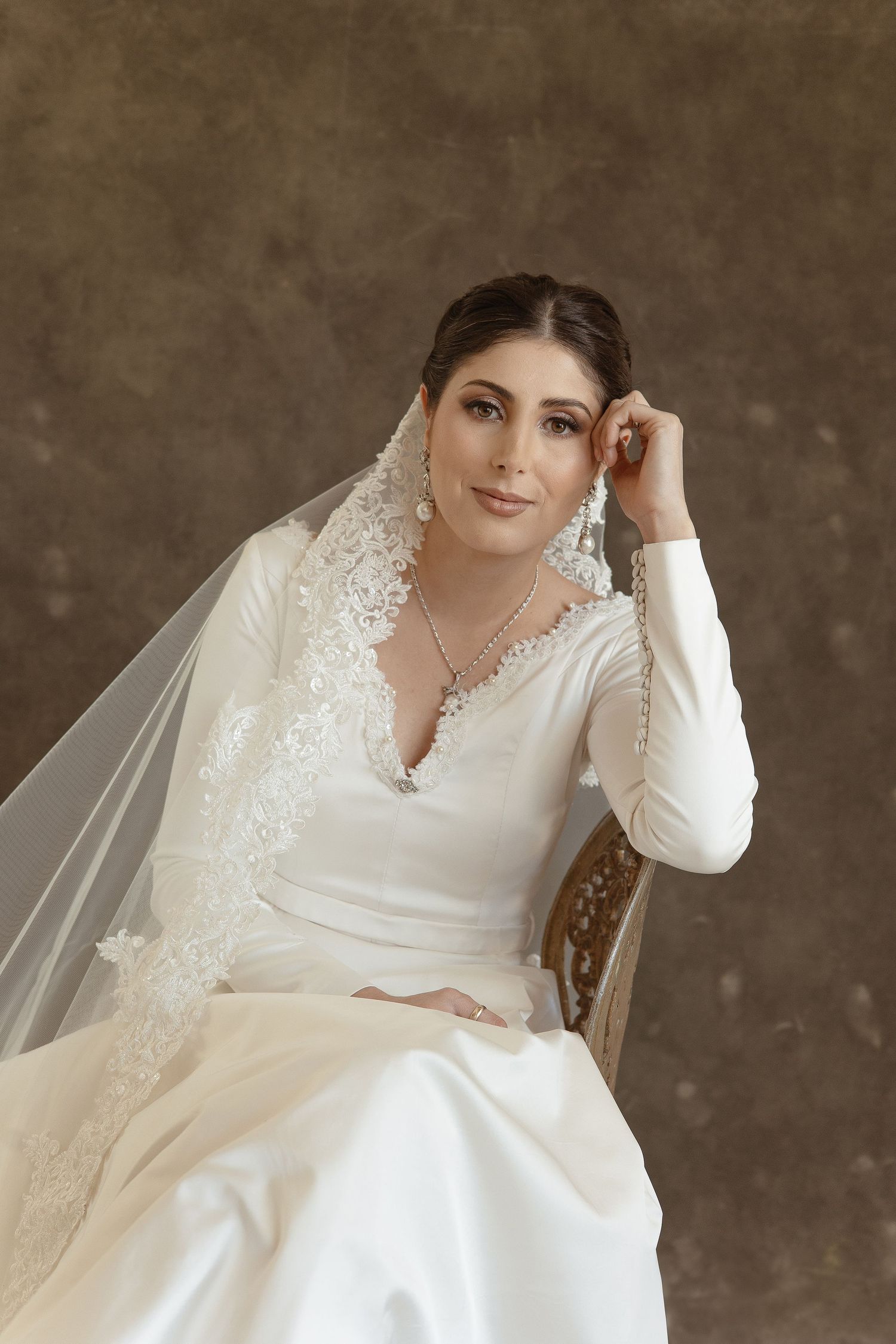 Elegant bridal portrait featuring a long-sleeved white wedding gown with lace details and flowing veil against a textured backdrop.