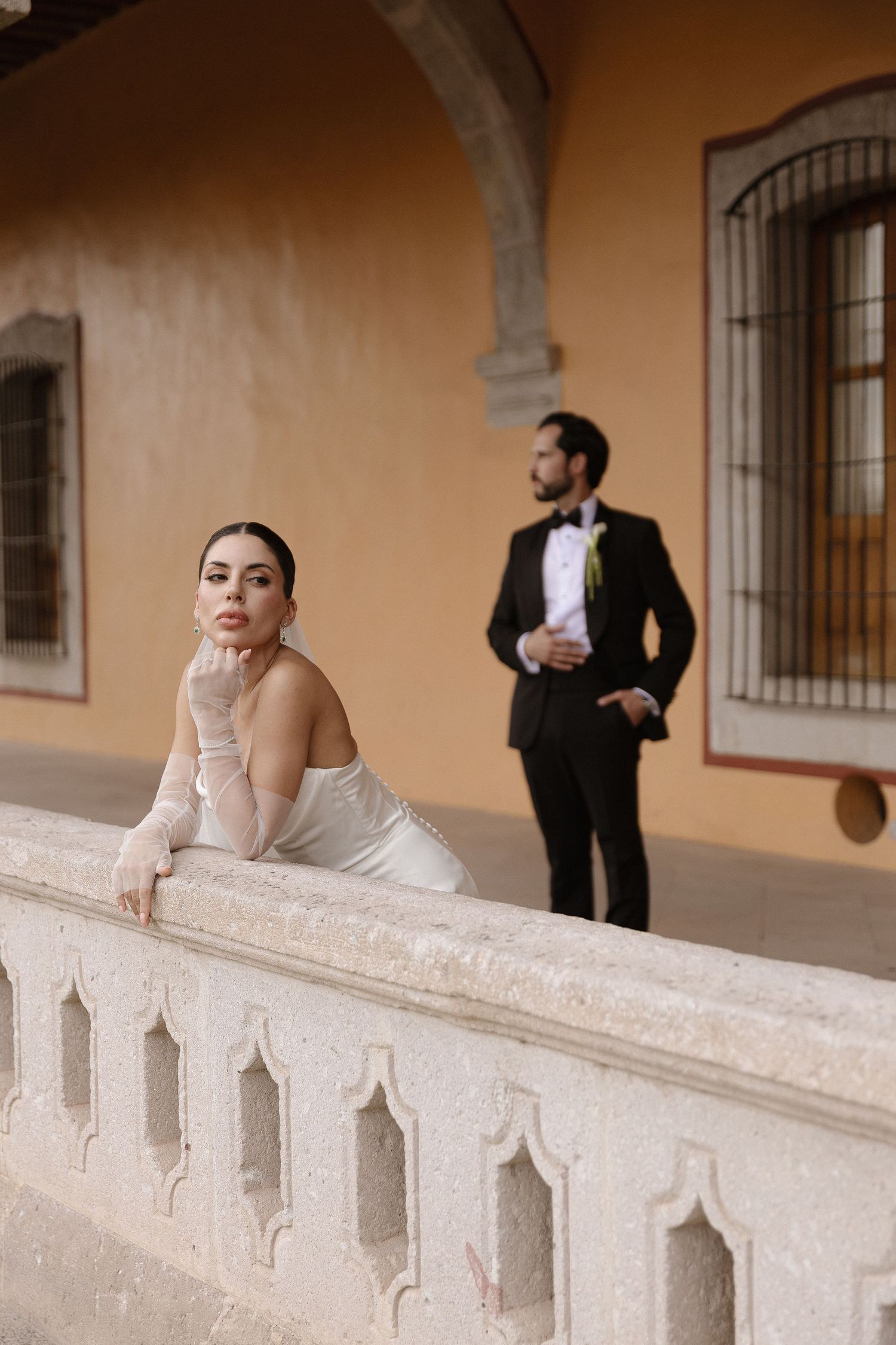 Elegant bride in white strapless gown leans on ornate stone balustrade while groom in black tuxedo stands behind her.