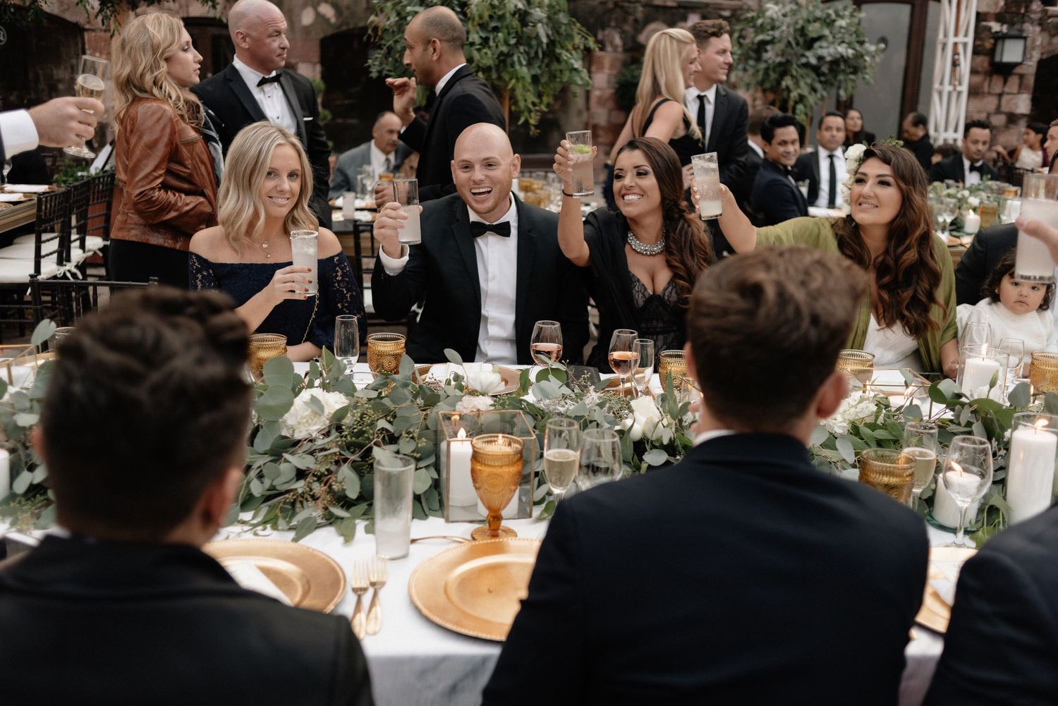 Guests seated at an elegantly decorated wedding reception table with greenery centerpieces and wine glasses.