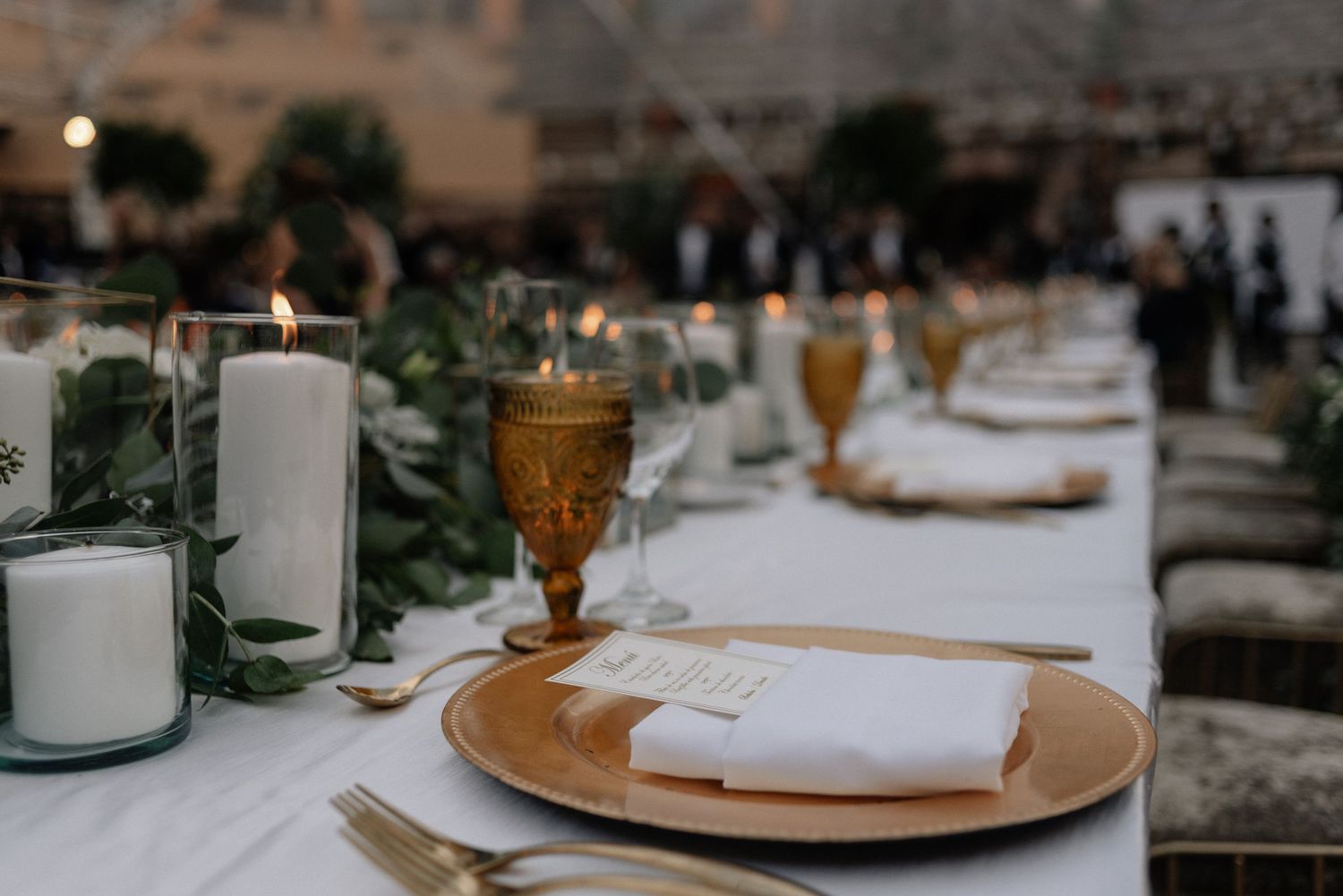Elegant wedding table setting with white candles, amber goblets, and place settings on a white tablecloth.