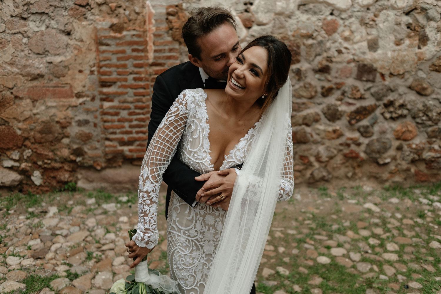 Newlywed couple shares a joyful moment together against rustic brick wall surrounded by autumn leaves.