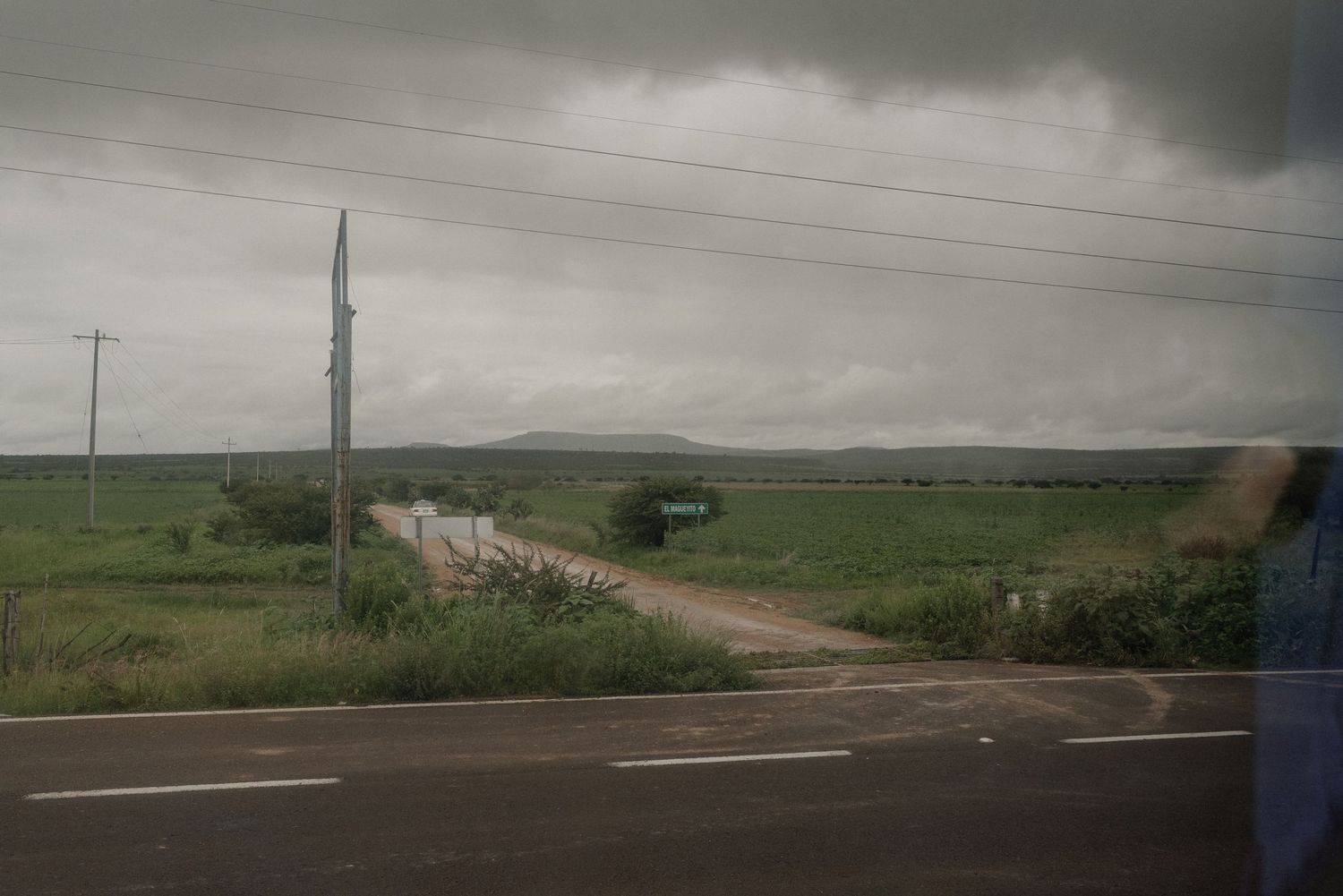 A dirt road extends into green farmland under stormy gray skies with utility poles lining the path.