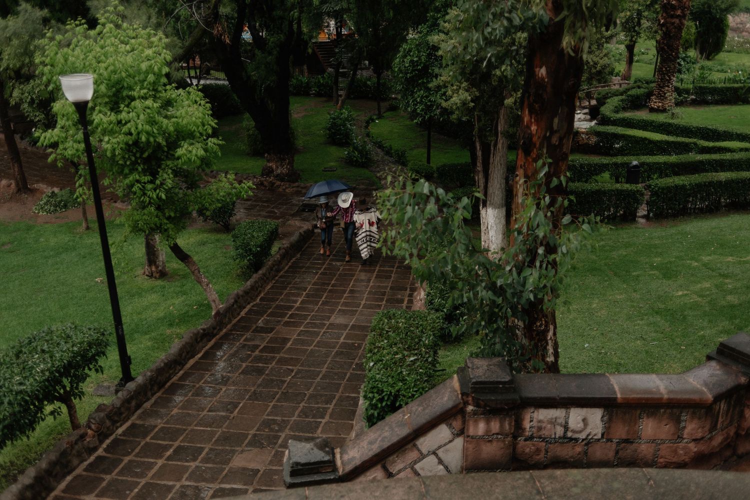 A stone pathway curves through a lush green garden with lamp posts and stone walls on a rainy day.