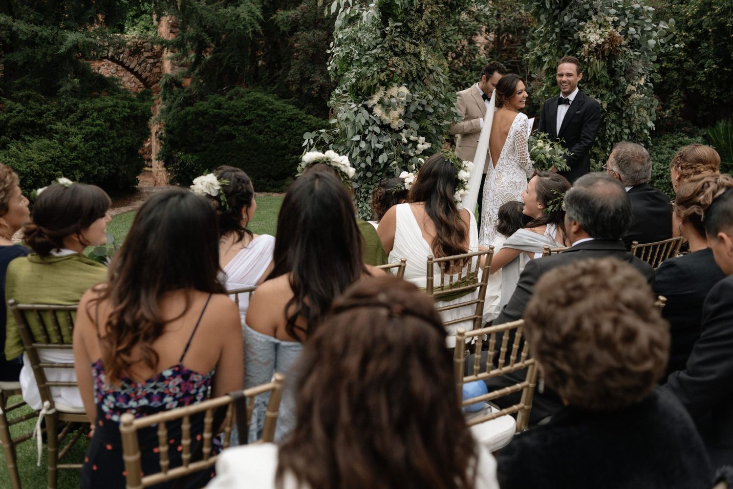 An outdoor wedding ceremony takes place in a lush garden setting with guests seated on gold chiavari chairs.