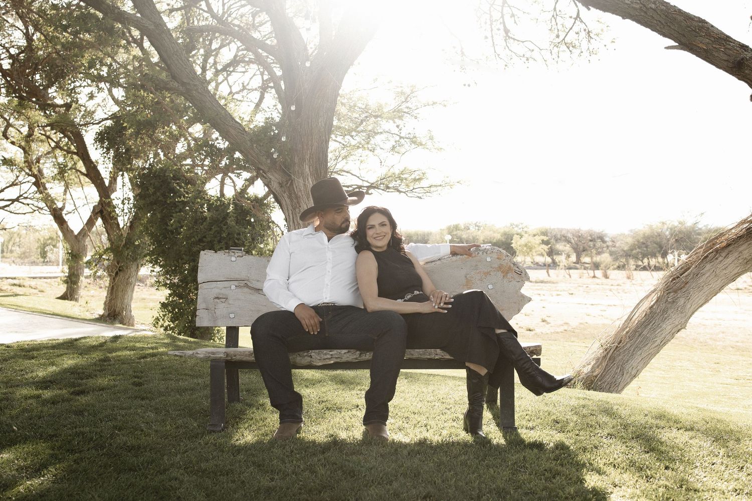 A couple sits together on a wooden bench under a tree during golden hour in a peaceful park setting.