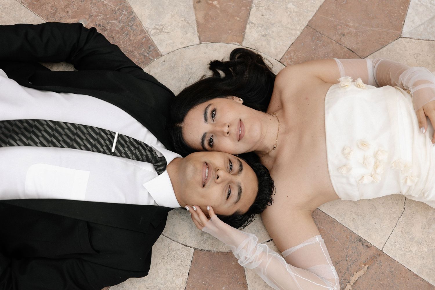 A couple in wedding attire lies on a tiled floor, with their heads together while smiling upward.