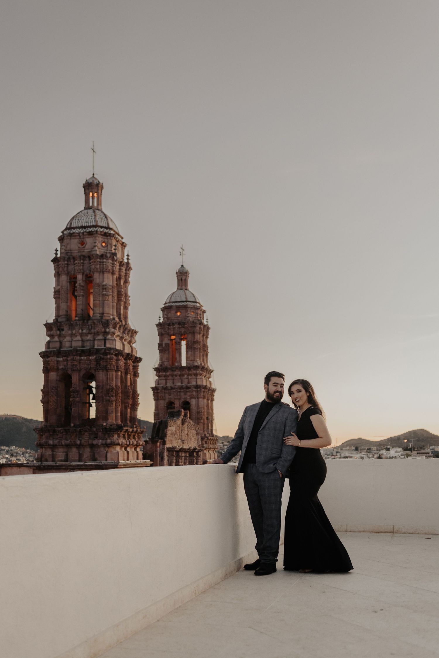 A couple stands against a backdrop of historic Spanish colonial church towers on a white rooftop.