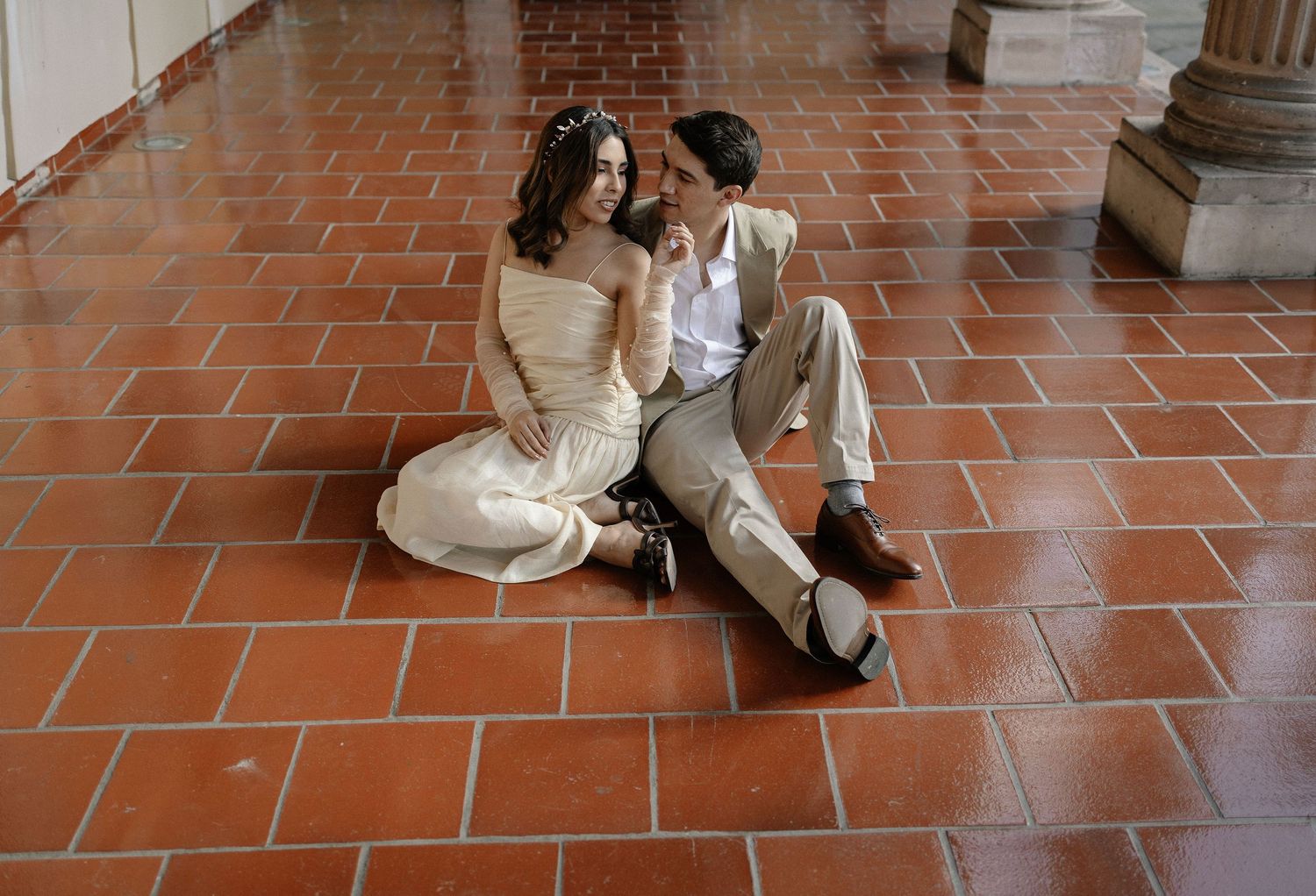 A couple in wedding attire sits intimately on terracotta tile flooring in a columned corridor.