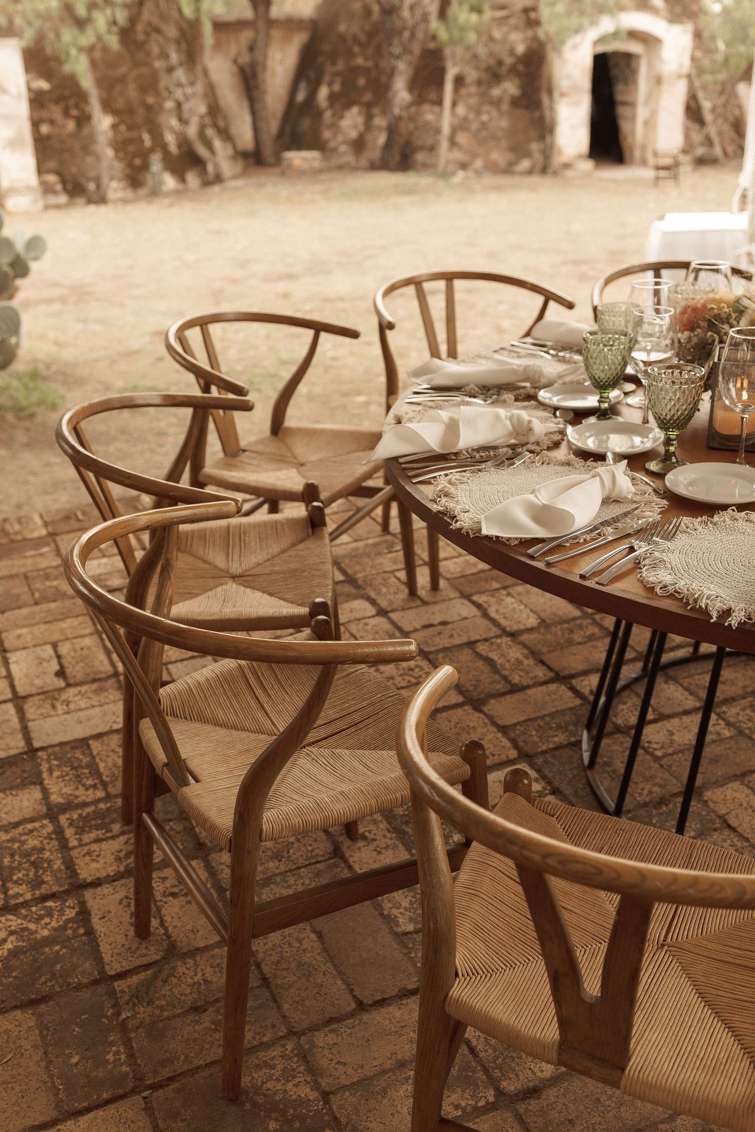 A series of wooden wishbone chairs with woven seats arranged on a stone patio in warm sepia tones.