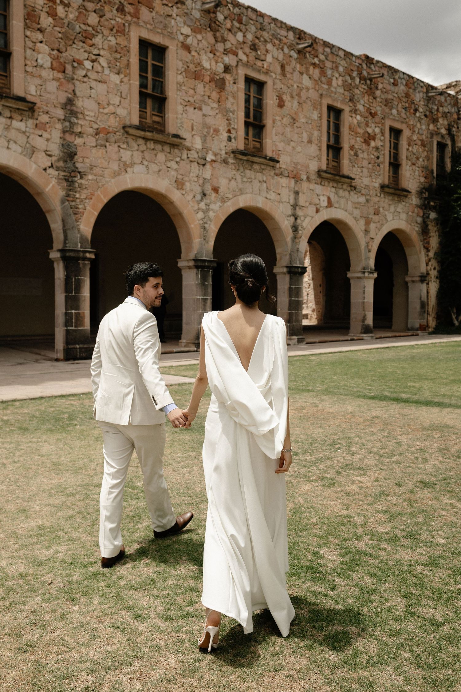 A couple in white outfits walking through an old stone archway courtyard with historic architecture.