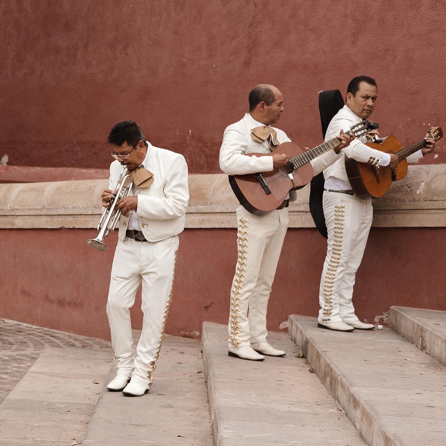 Three mariachi musicians in white uniforms perform with their instruments against a terracotta wall.