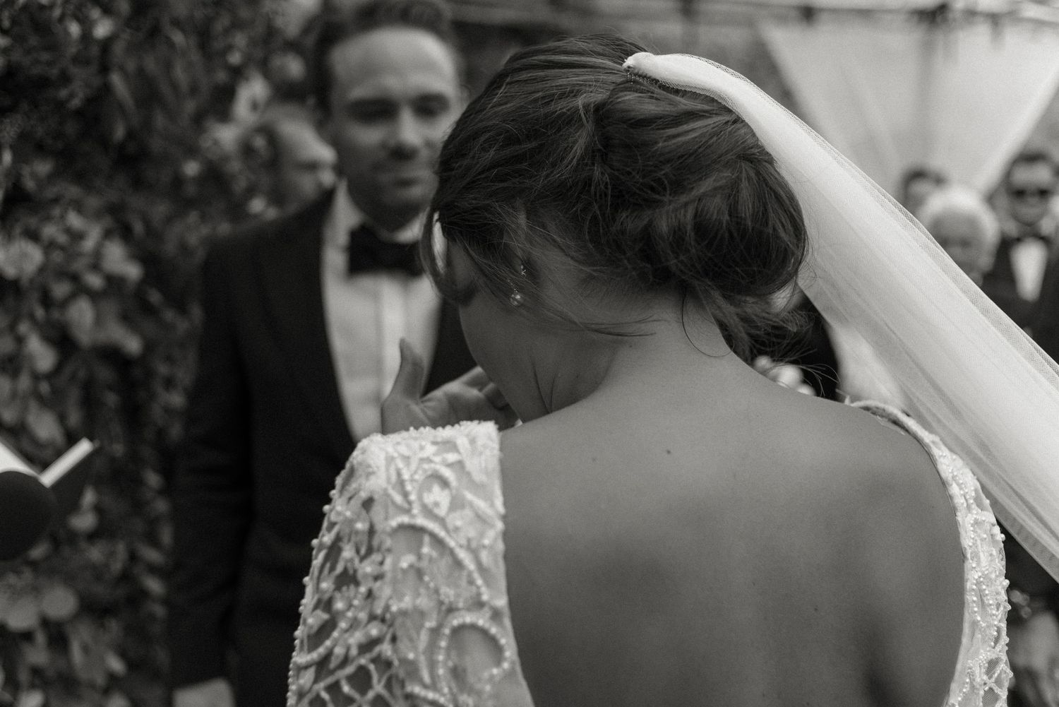 Black and white wedding photograph showing bride wearing lace dress with open back detail as groom looks on in background.