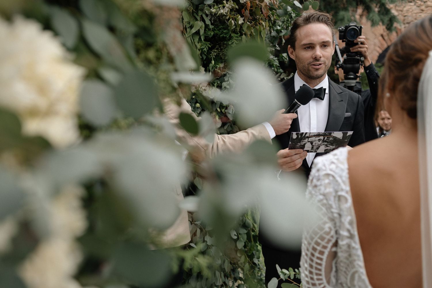 Groom in a black tuxedo and bowtie stands among white flowers and greenery during an outdoor wedding ceremony.