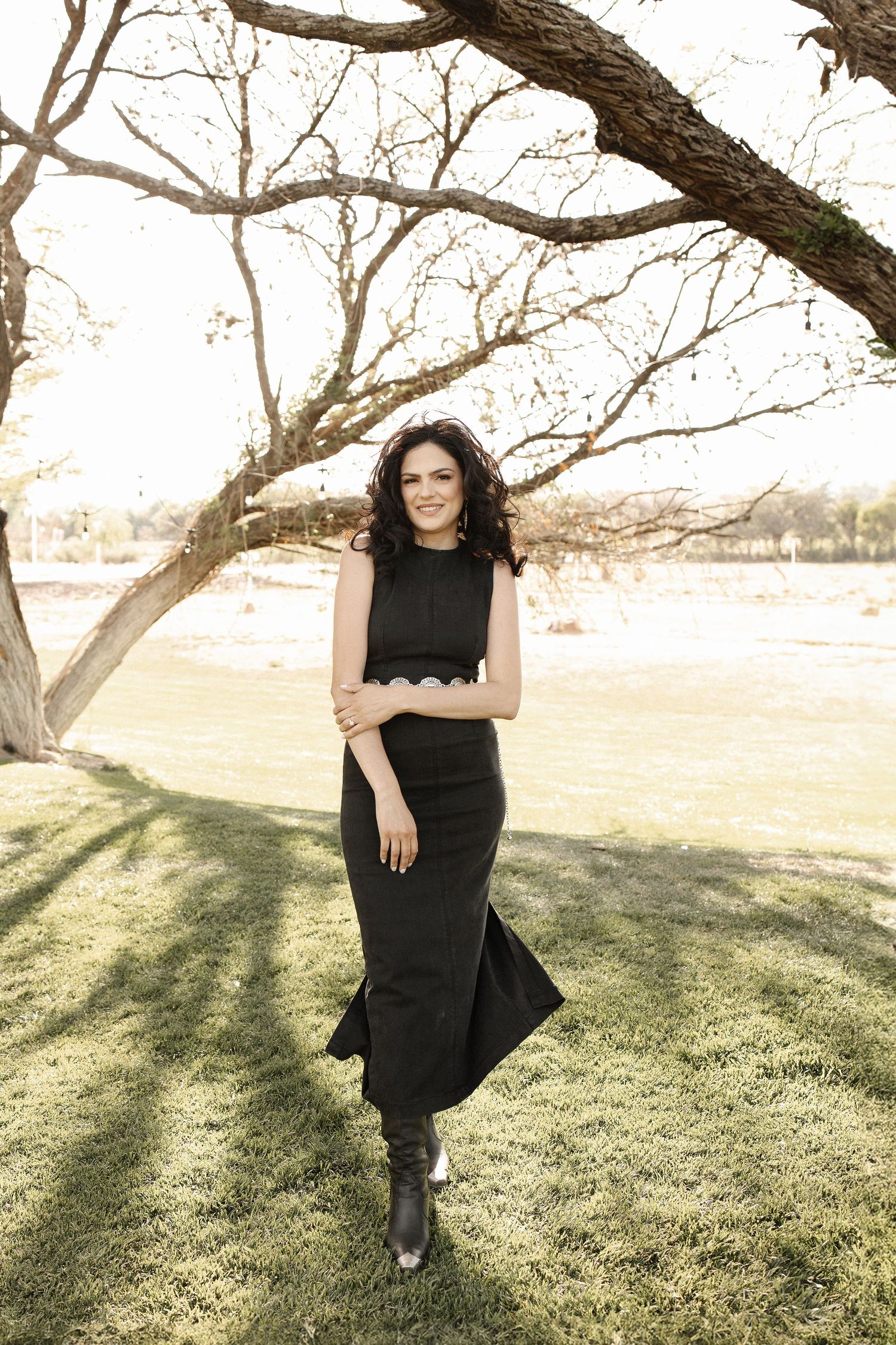 A person in a black dress poses outdoors near a tree on a grassy lawn during golden hour.