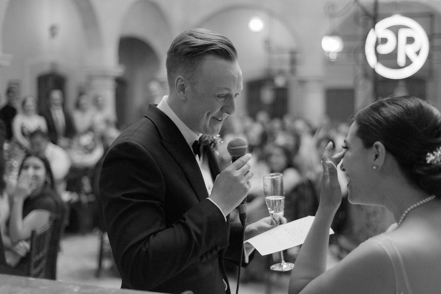 A black and white reception moment captured during wedding speeches with guests in the background.