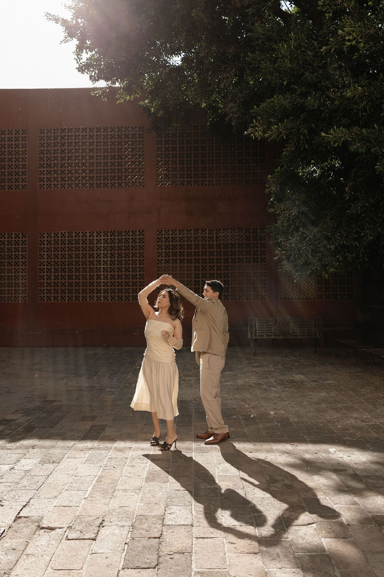 Couple dancing together on a sunlit stone patio casting dramatic shadows against a brick wall.