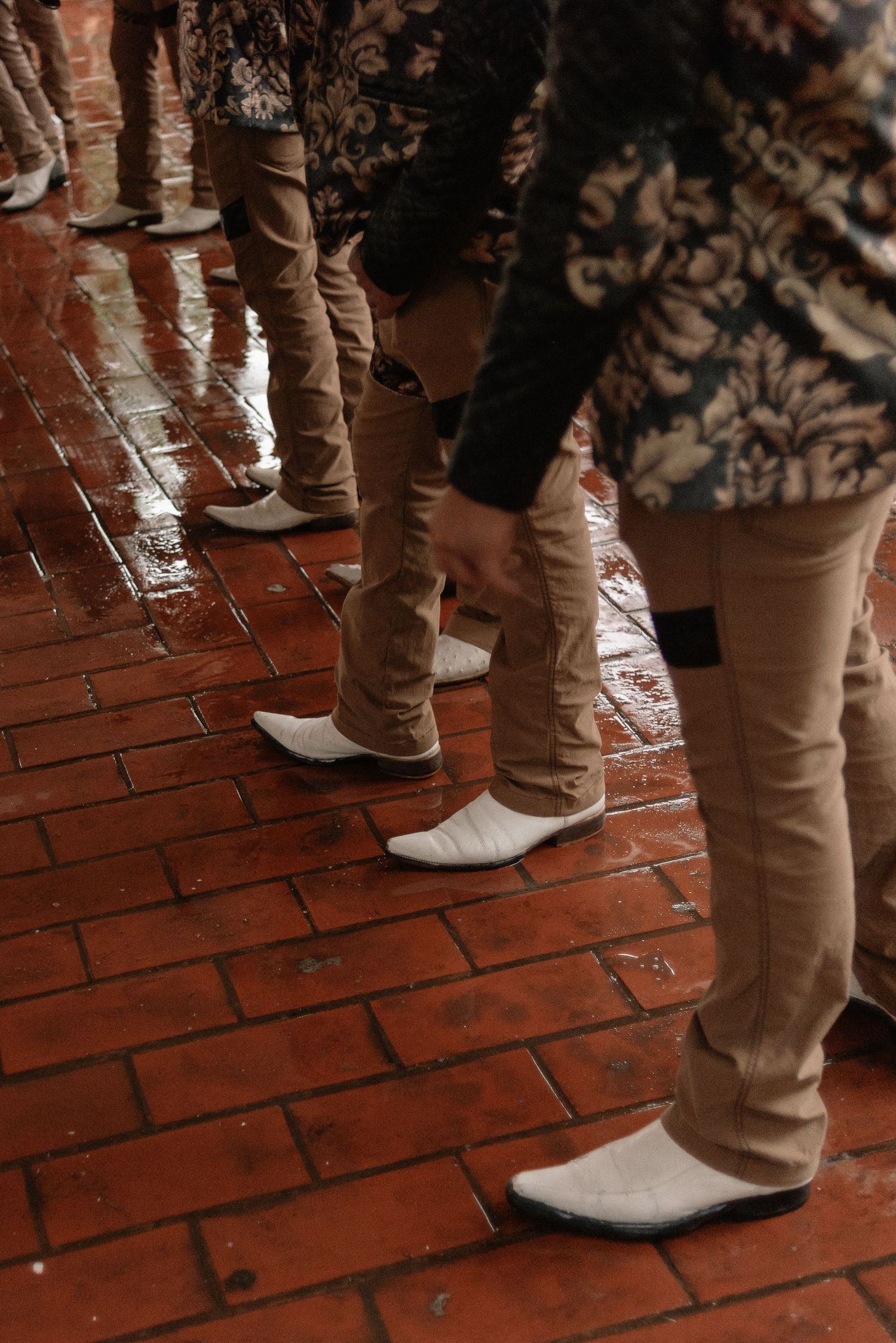 Row of people wearing khaki pants and white cowboy boots standing on wet red brick pavement in warm lighting.