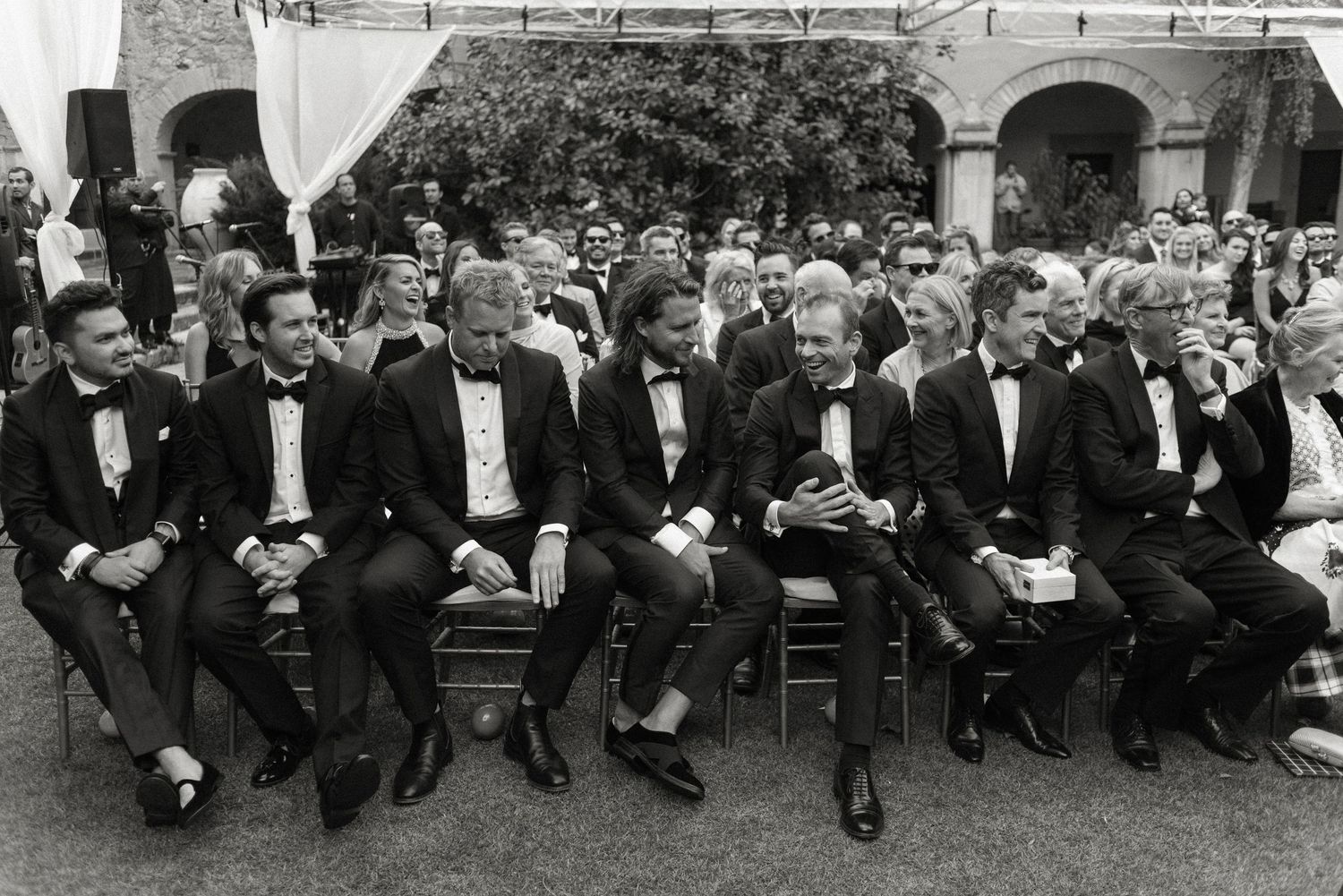 A group of men in formal black tuxedos and bow ties sit in the front row at an outdoor event.