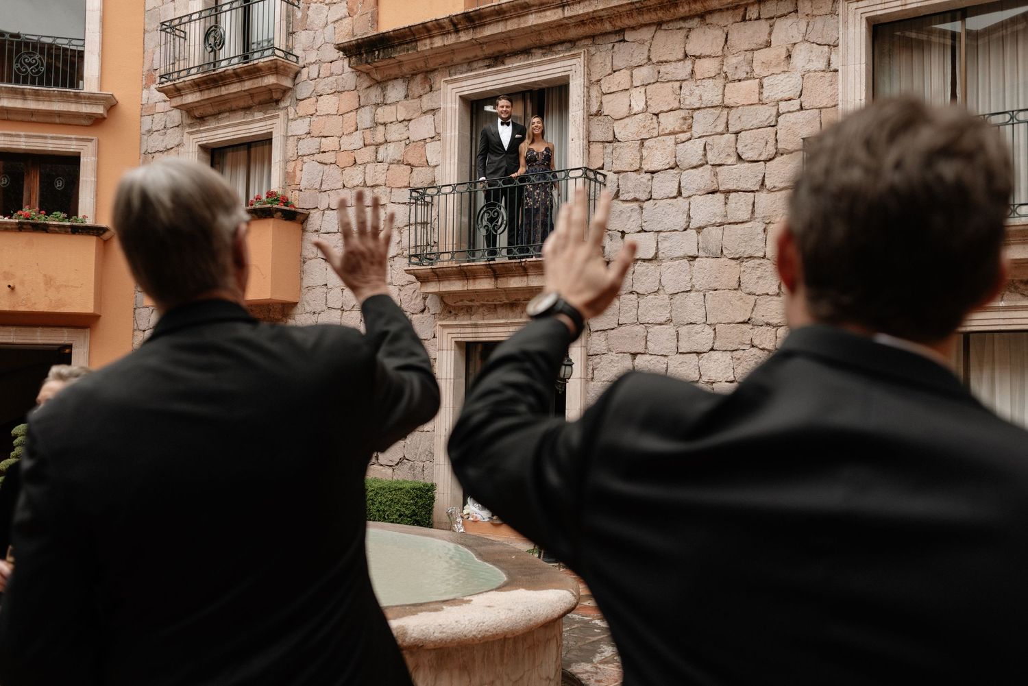Two people in black suits wave up at a couple standing on a stone balcony during a celebration moment.