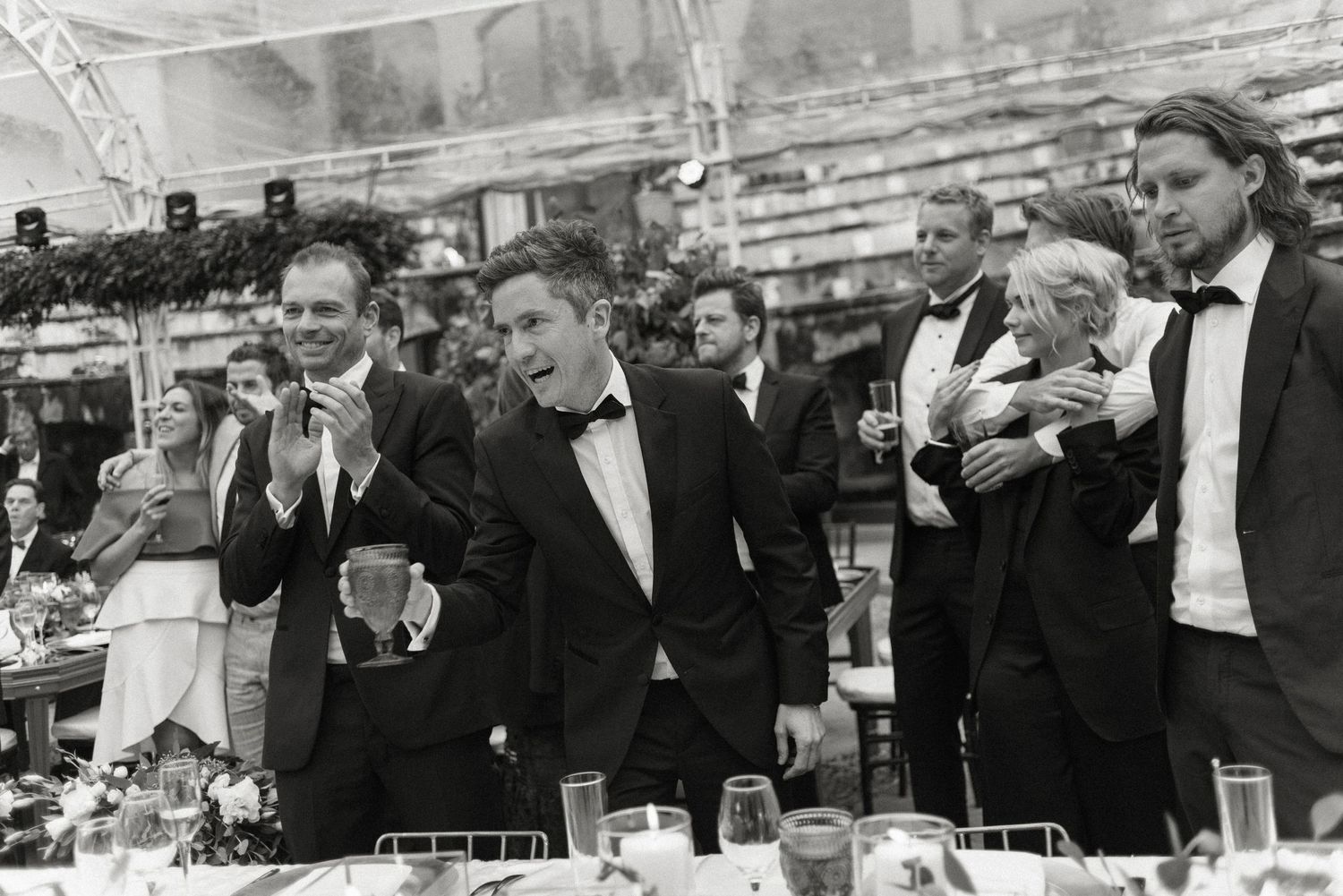 A black and white photo shows suited business people laughing and having drinks at an elegant social gathering.