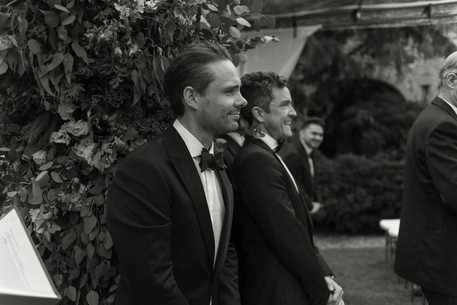 A black and white photograph shows wedding guests in tuxedos at a formal outdoor ceremony.