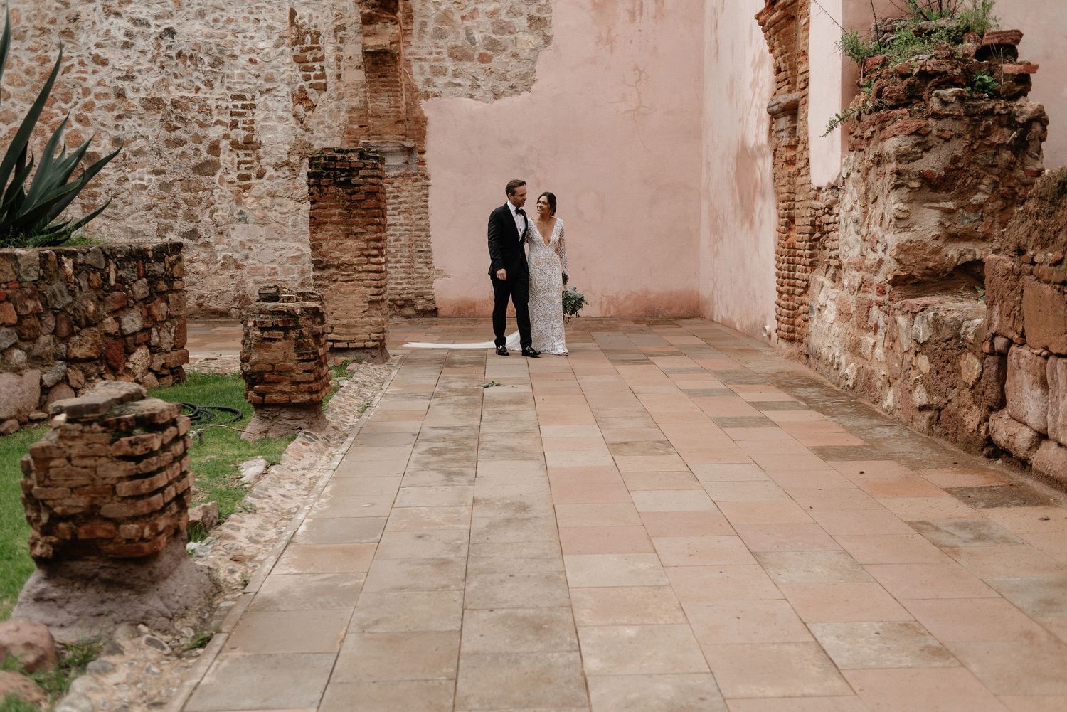 A couple stands in a rustic courtyard with stone walls and tiled floors at a historic architectural site.