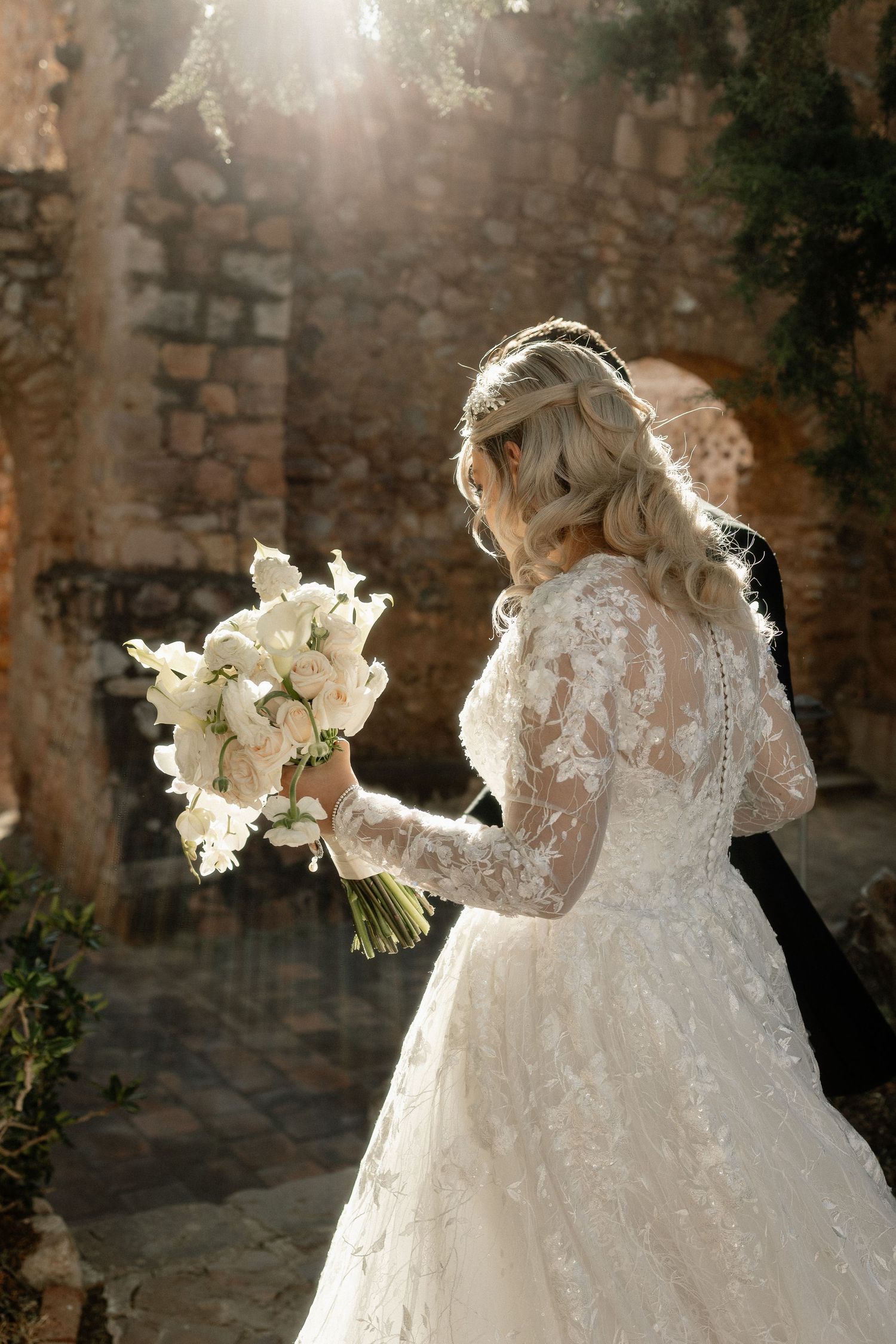 A romantic wedding photo series showing a bride in a lace gown holding a white floral bouquet against a rustic stone wall.