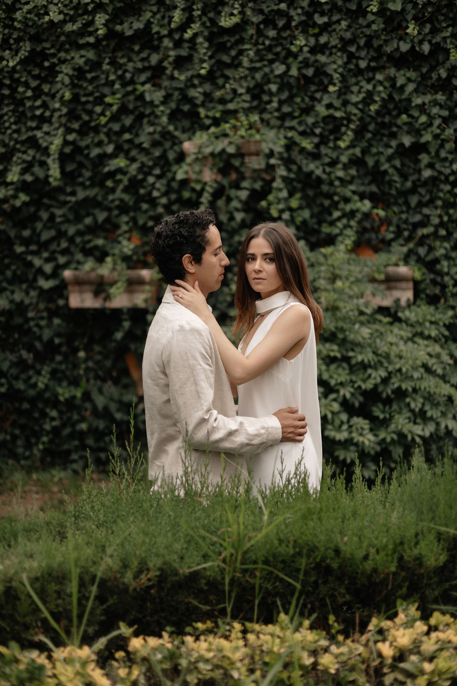A couple embraces in front of an ivy-covered wall in a romantic garden setting.