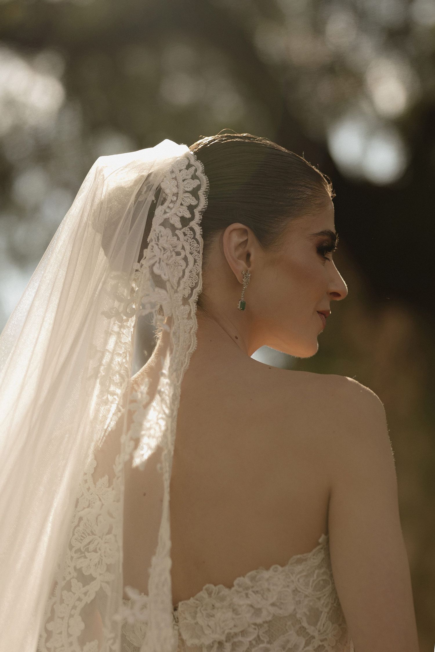 A bride in a strapless wedding dress is shown in profile with delicate lace veil details against a soft bokeh background.
