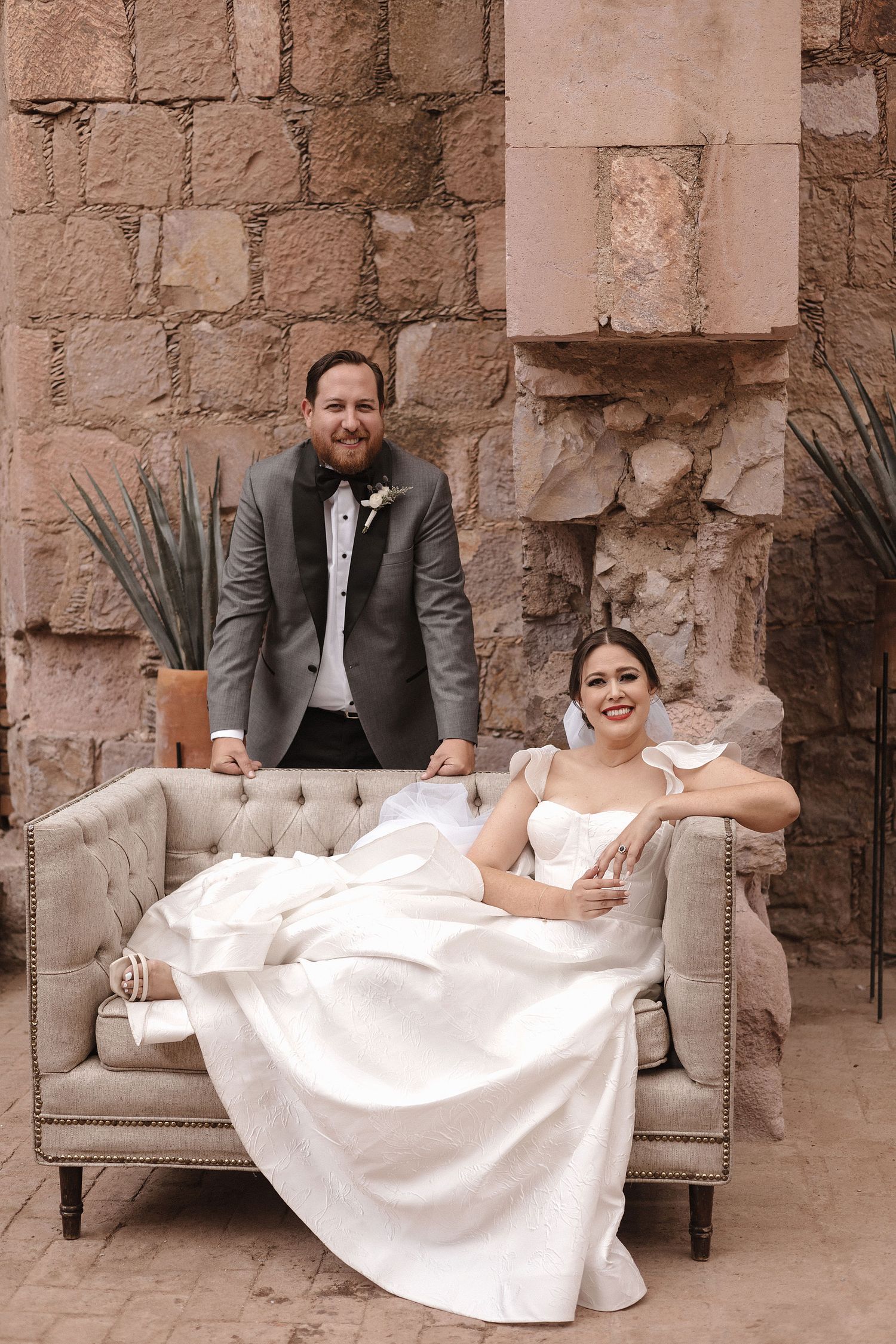 Wedding couple posing on vintage sofa against rustic stone wall with desert plants.