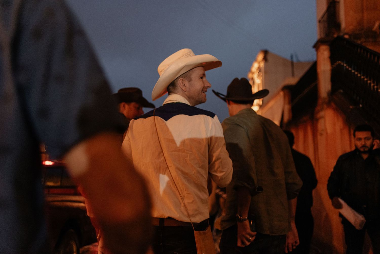 Person wearing a cowboy hat and two-tone jacket stands among a small crowd at dusk on a city street.