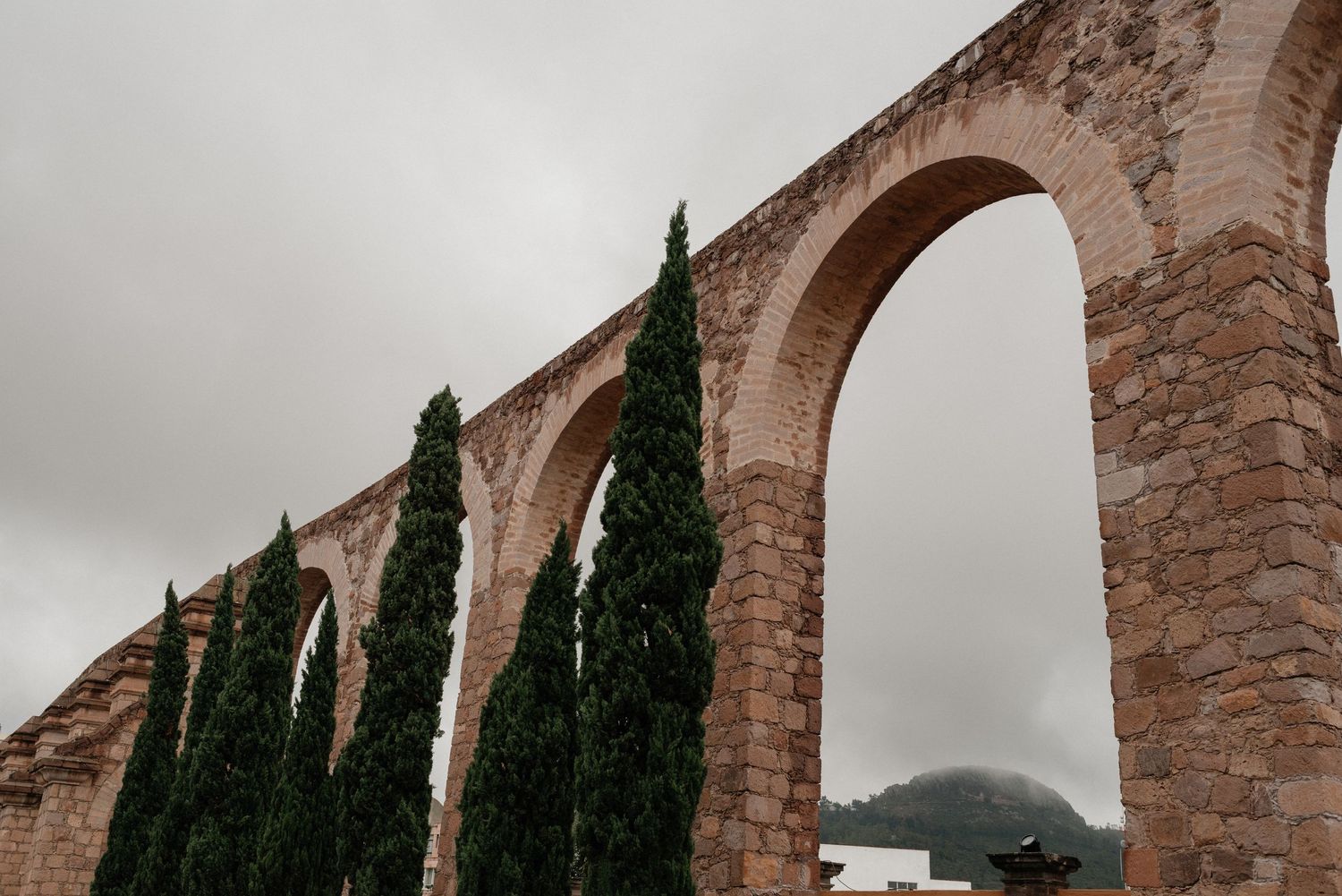 Ancient stone aqueduct with tall cypress trees growing along its arches against a misty mountain backdrop.