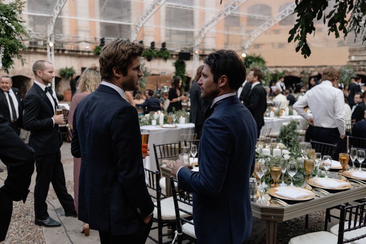 Two guests in navy suits chat during an elegant outdoor wedding reception with rustic table settings and greenery decor.