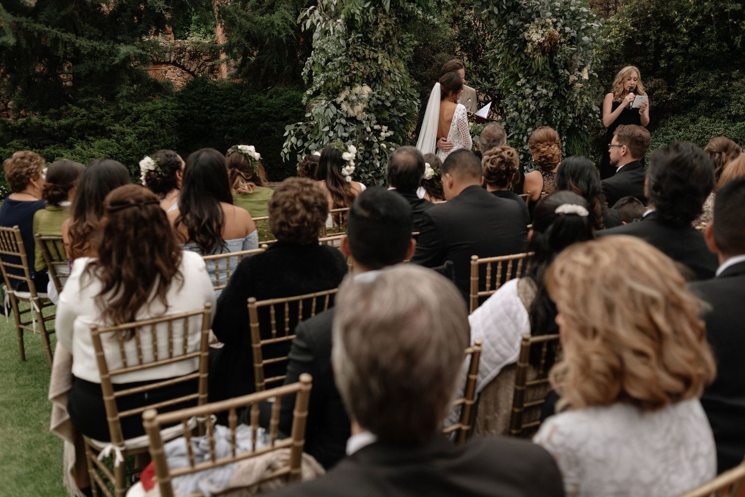 Wedding guests seated in gold Chiavari chairs watch an outdoor garden ceremony under trees and greenery.