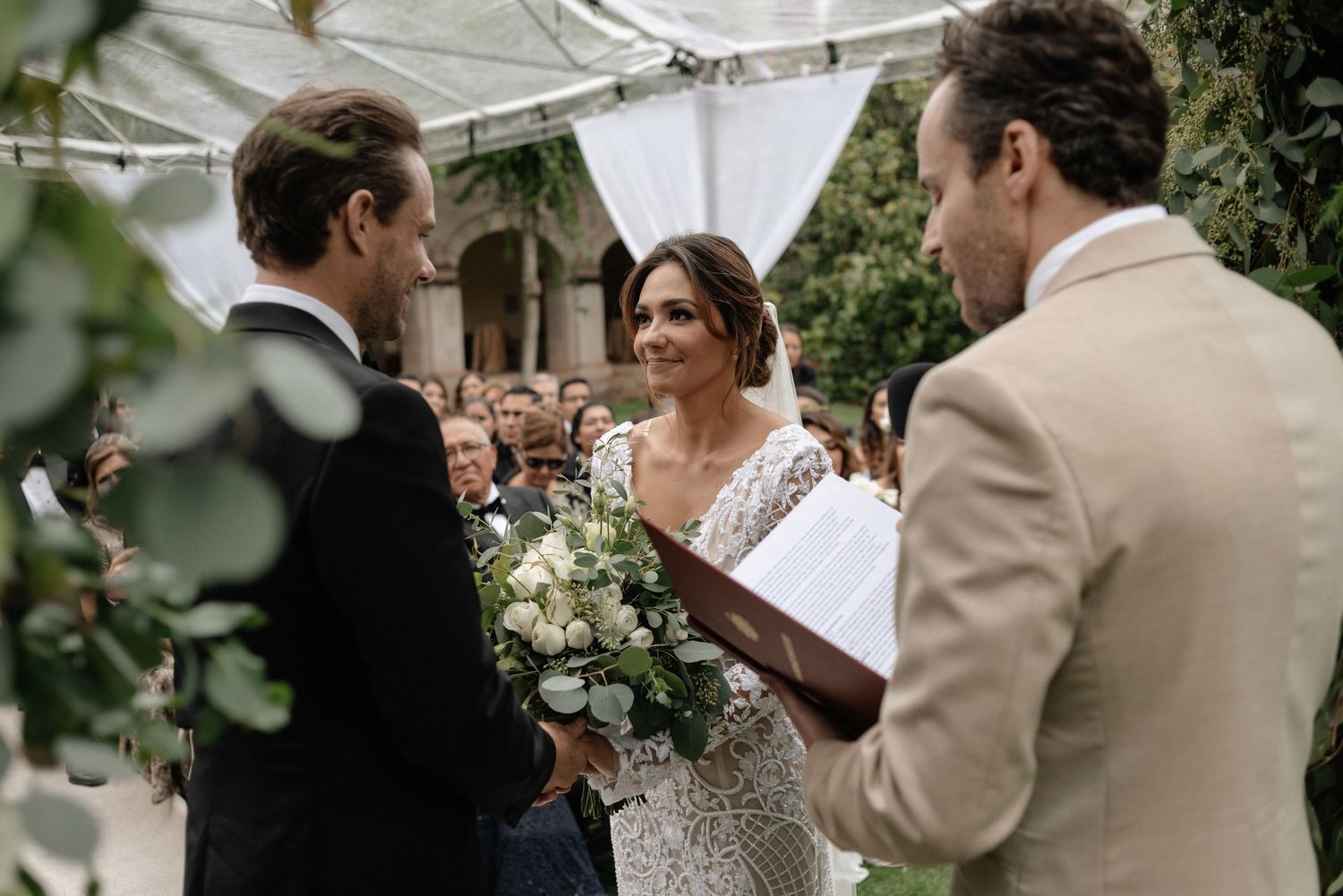 A bride and groom exchange vows during an outdoor wedding ceremony under a white tent canopy.