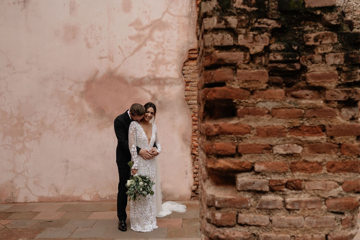 A couple shares a romantic moment next to an exposed brick wall and textured pink plaster surface.